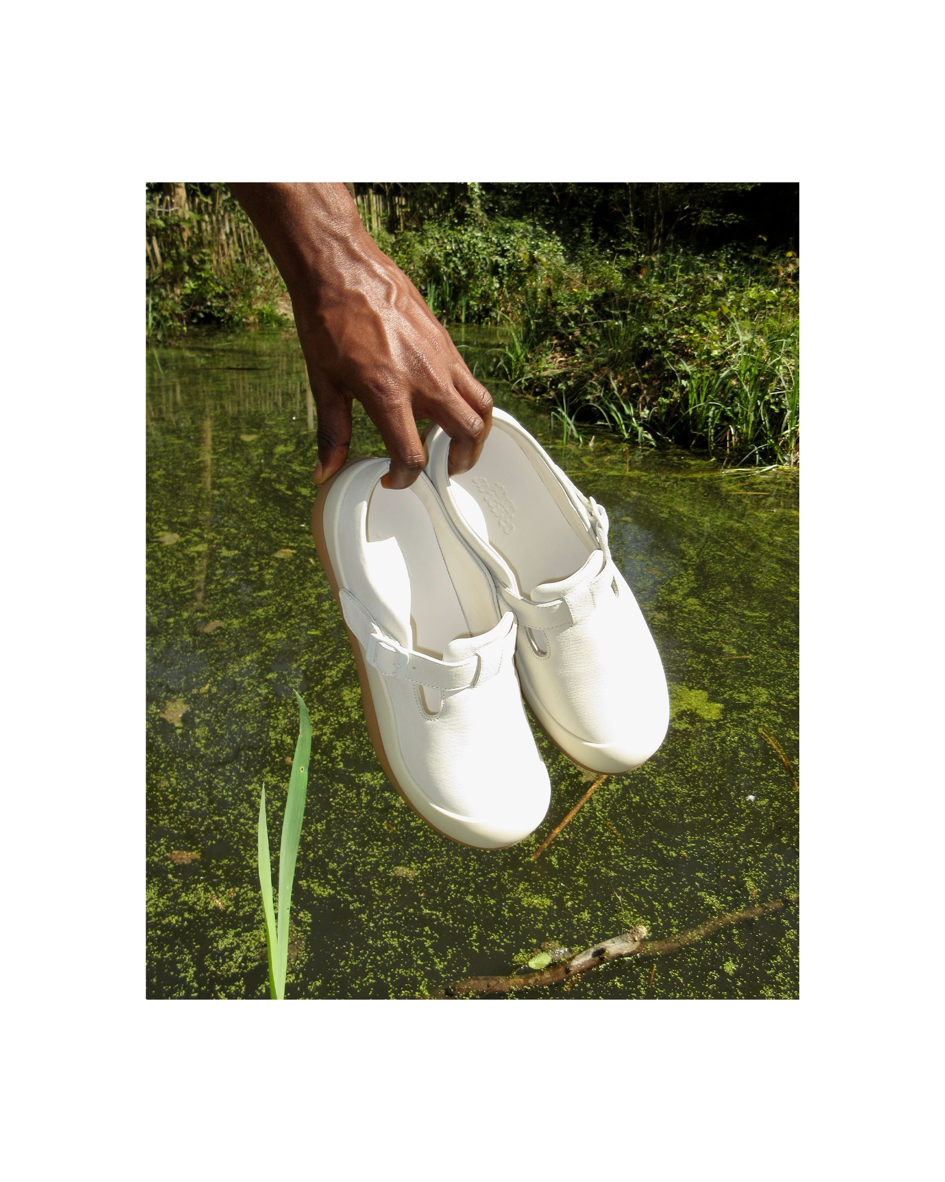 A hand holds a pair of white shoes above a lush, green pond with foliage in the background.