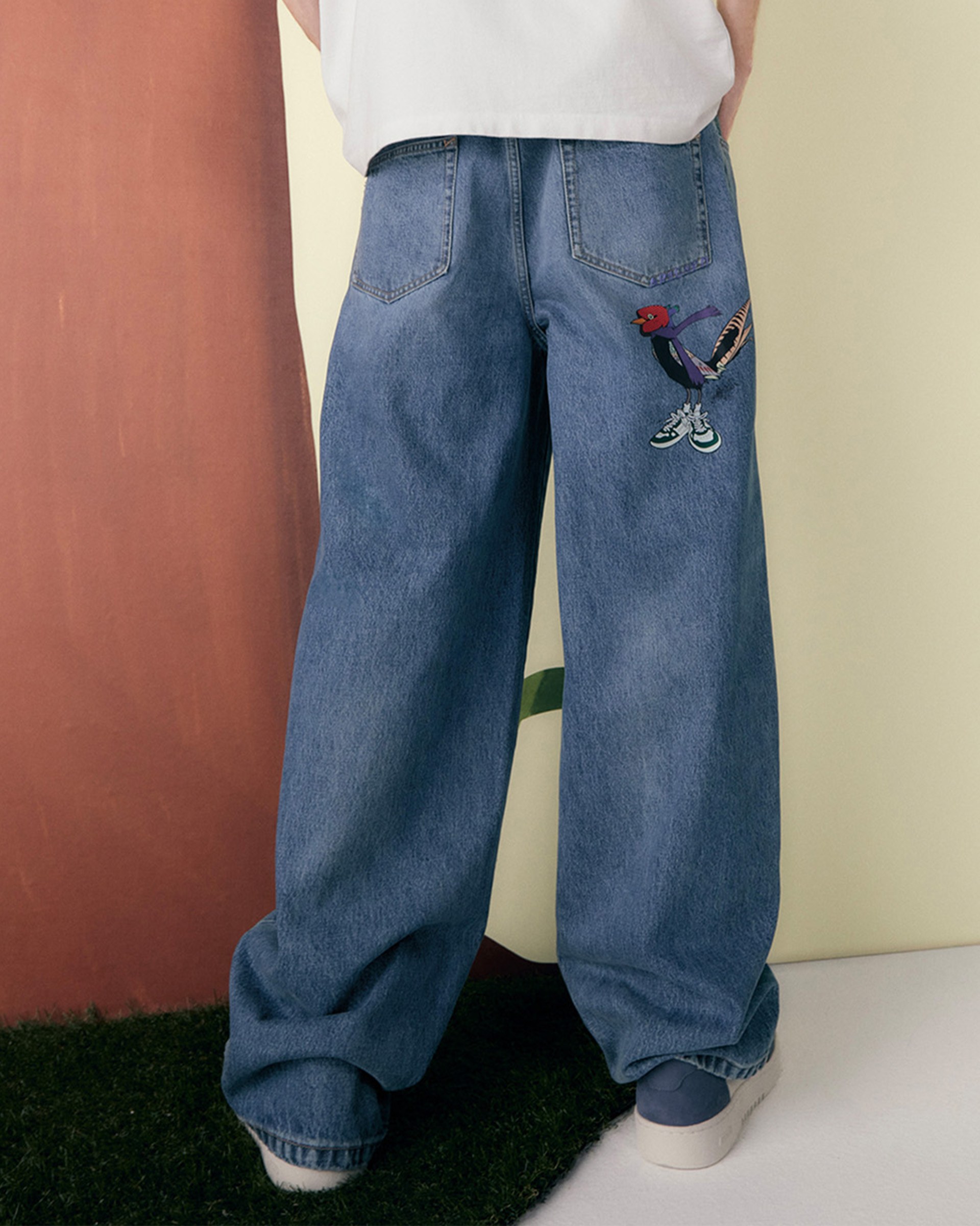 Person wearing wide-leg jeans with colorful embroidery on the back pocket, standing on grass with a white and brown backdrop.