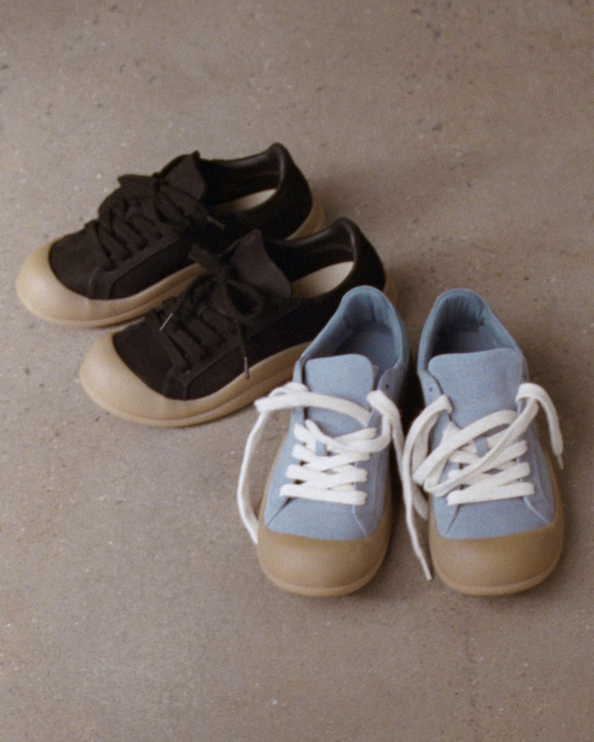 Two pairs of sneakers on a concrete floor: one black with dark laces, the other light blue with white laces, both with beige soles.