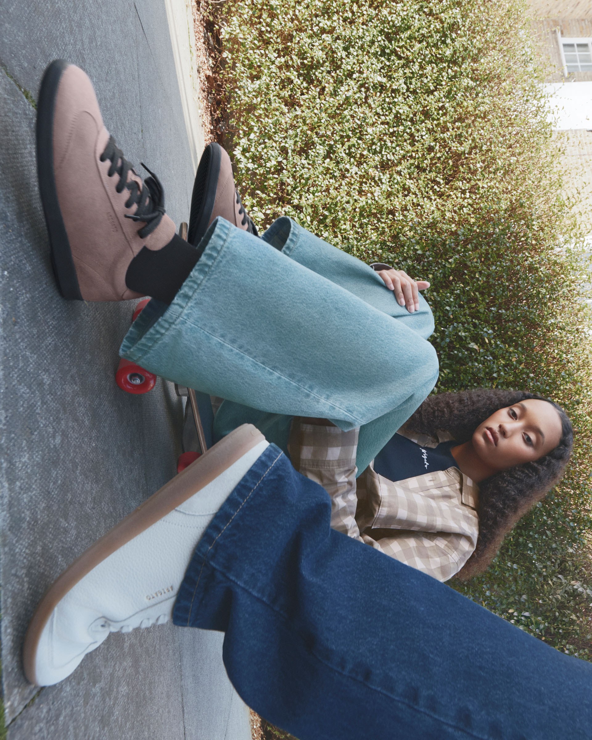 Person sitting on a skateboard wearing light jeans and brown shoes, next to another person in white shoes on a paved walkway with bushes.