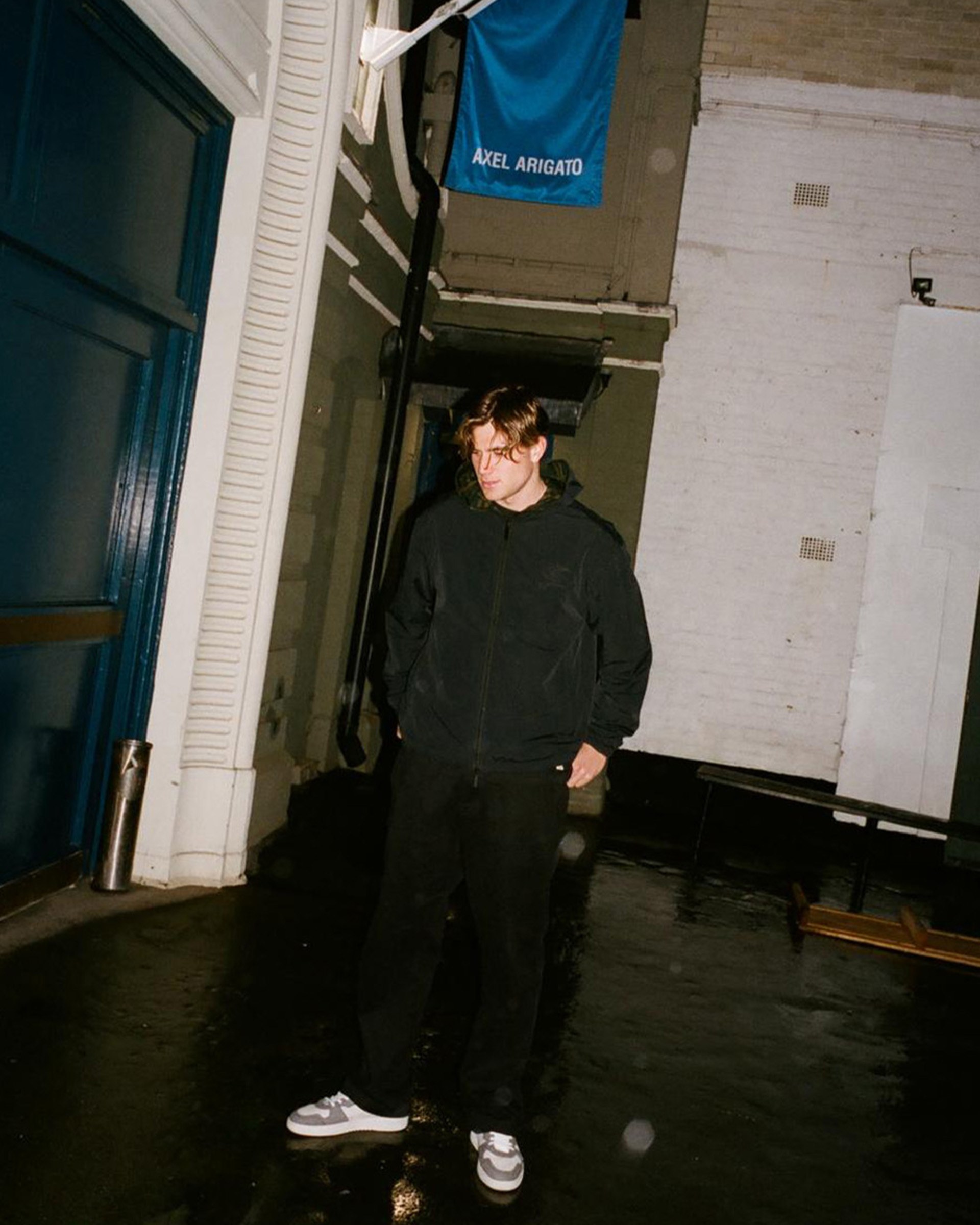 Person standing in a dimly lit urban alley, wearing dark clothing. A blue flag with the text "Axel Arigato" hangs above.
