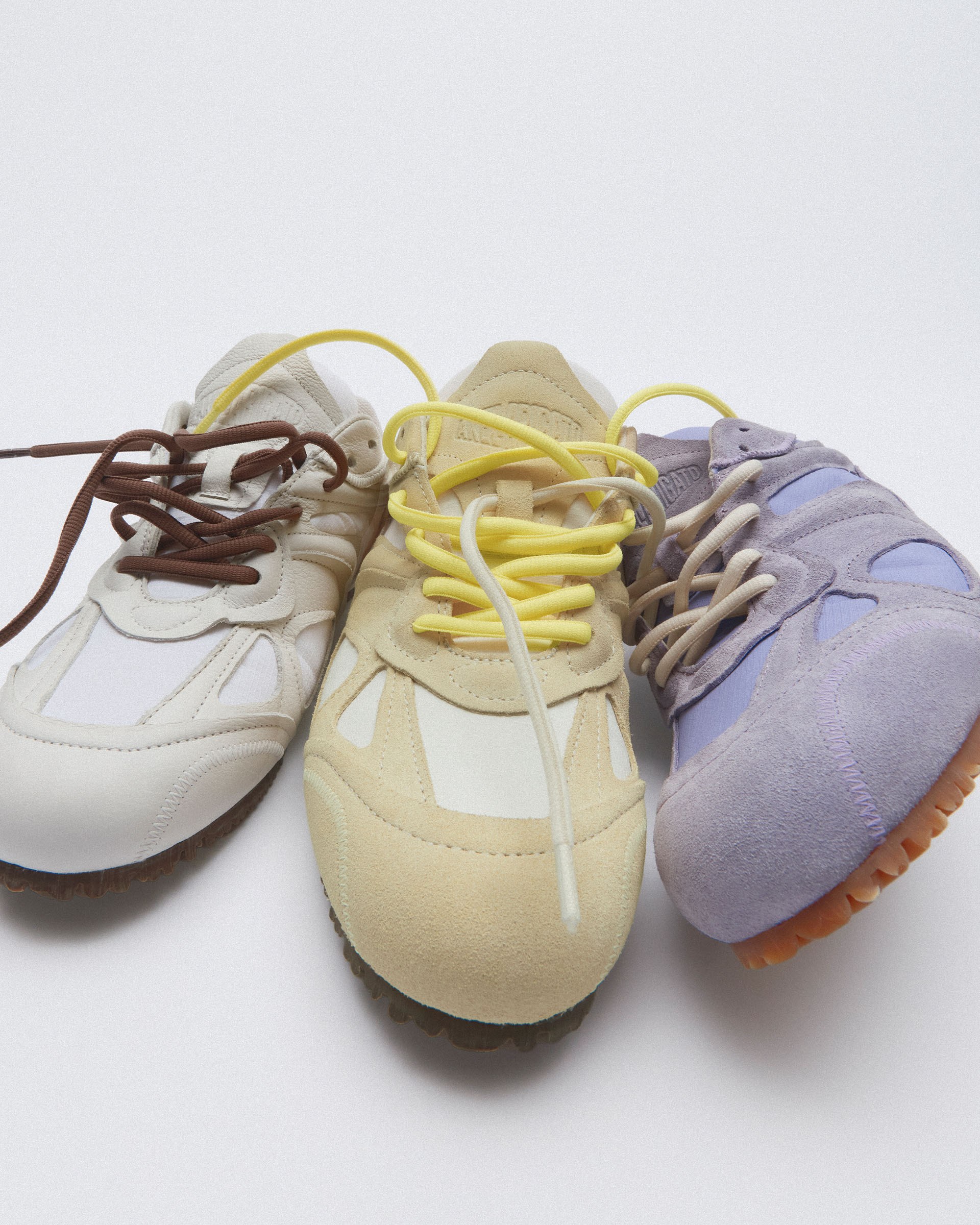 Three sneakers in beige, yellow, and lavender hues with contrasting laces, arranged side by side on a white background.