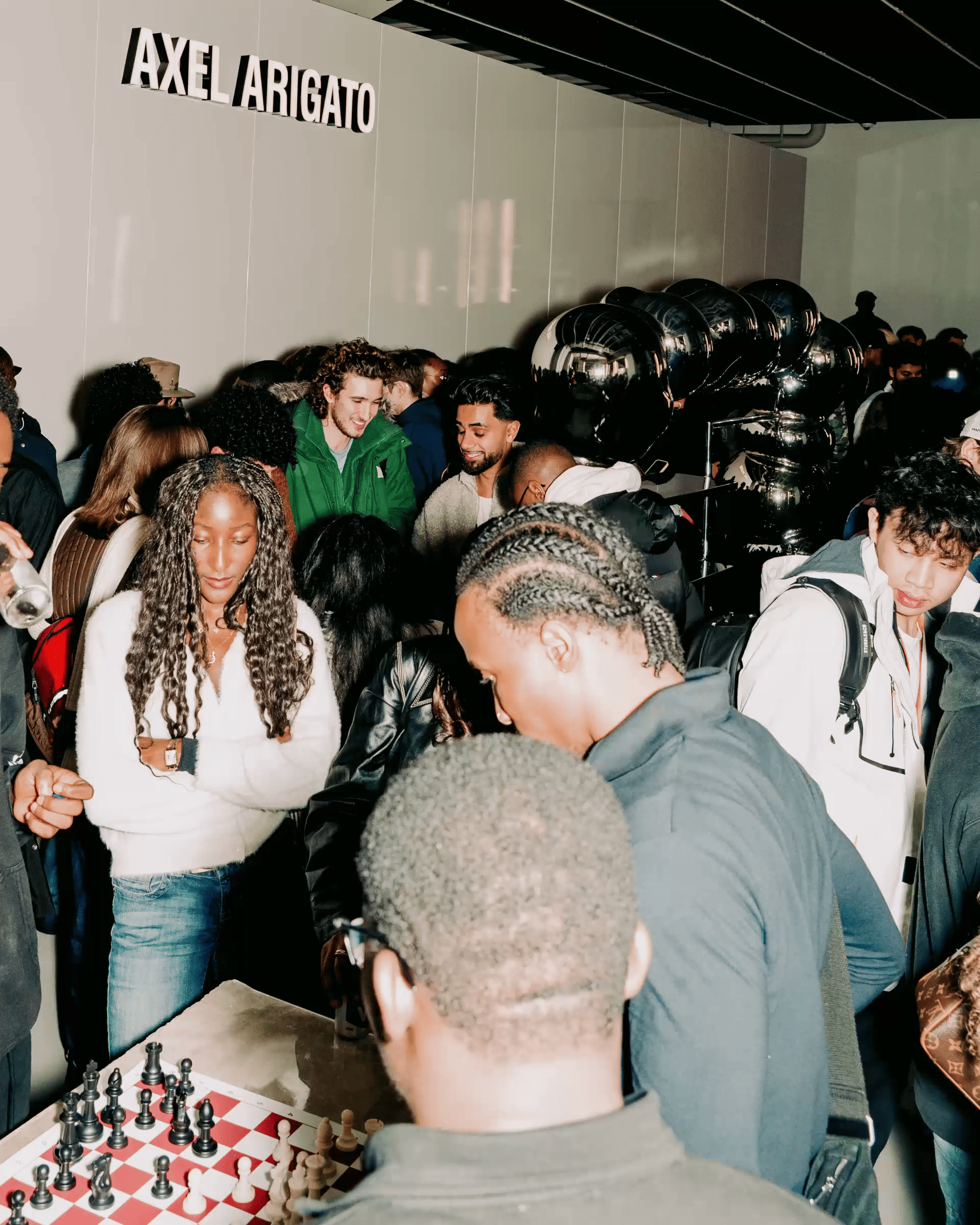 Crowded indoor space with people engaging in conversation and playing chess, under a sign that reads "AXEL ARIGATO."