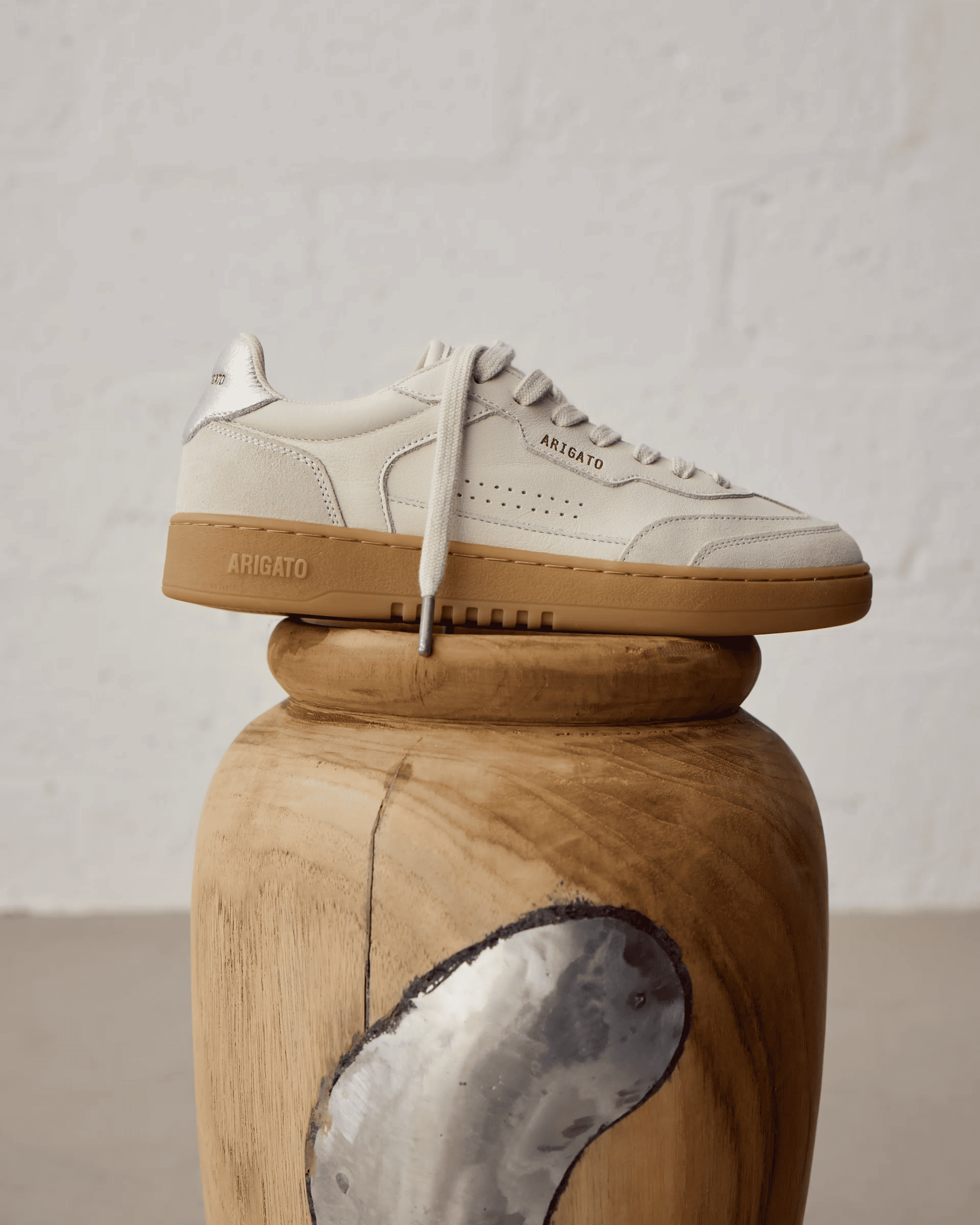 White sneaker with brown sole displayed on a wooden sculpture, against a textured white wall background.