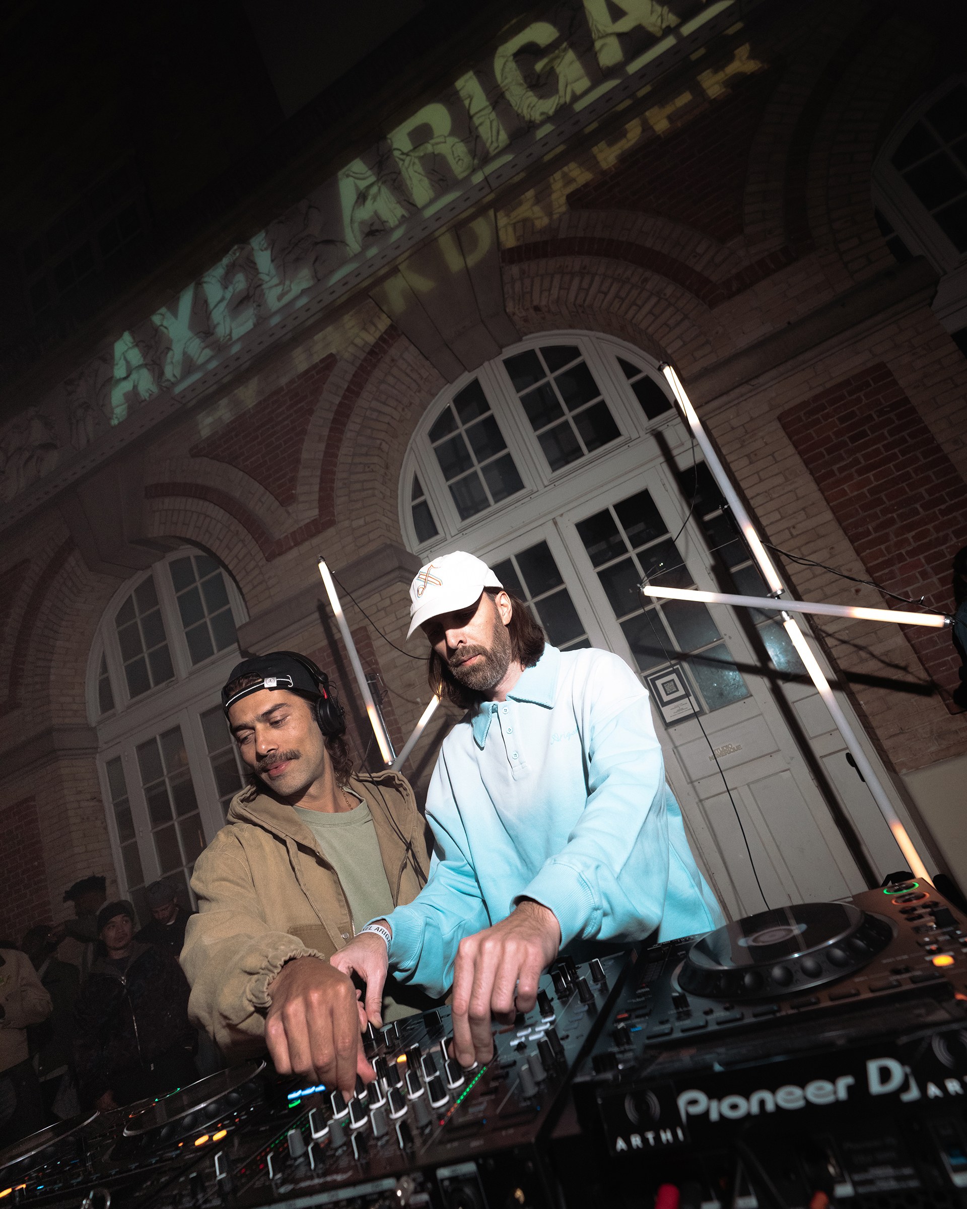 Two DJs perform together, mixing tracks on a soundboard, with "AXEL ARIGATO" projected on a building wall behind them.