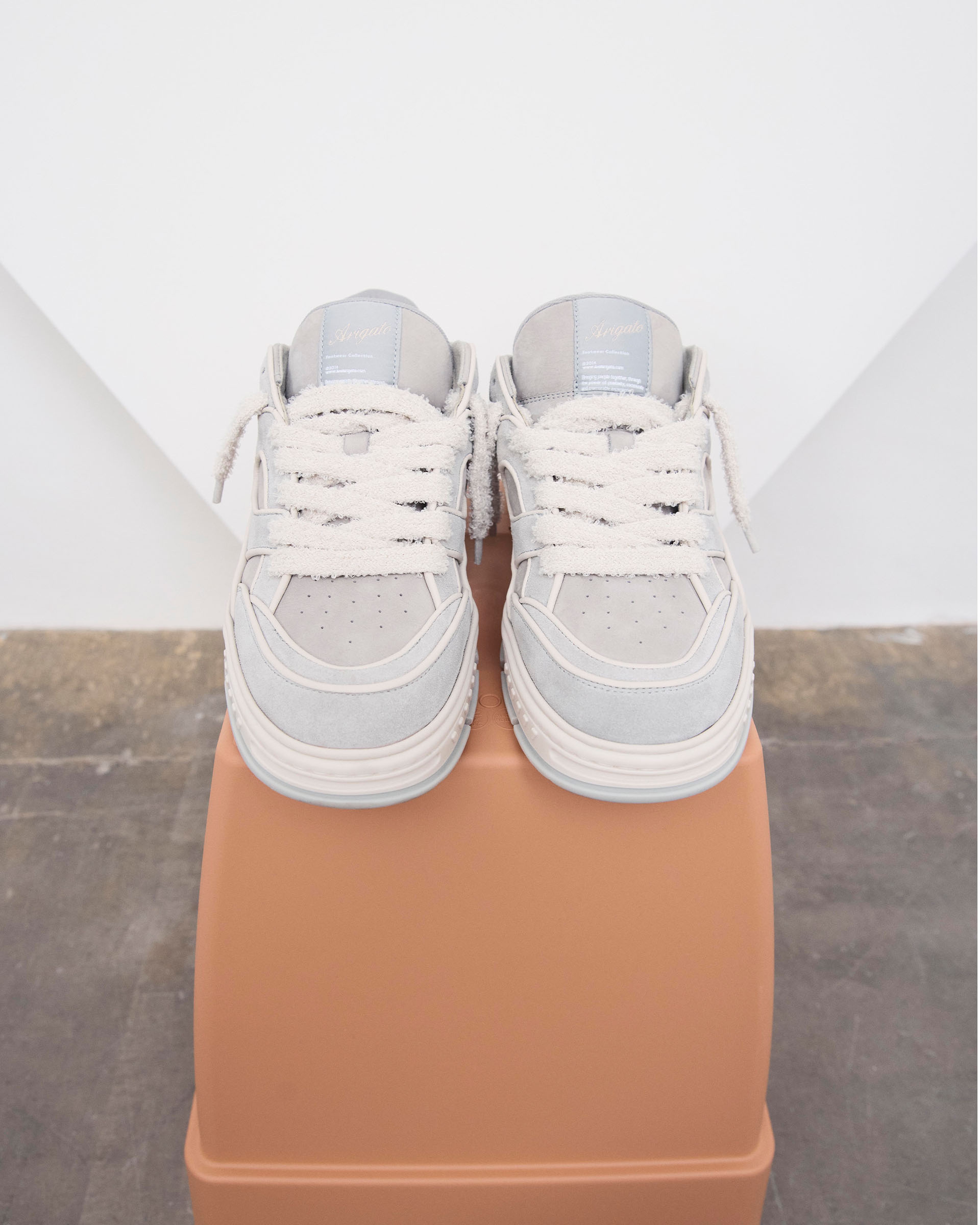 A pair of white sneakers with thick laces displayed on a rectangular, brown platform against a neutral background.