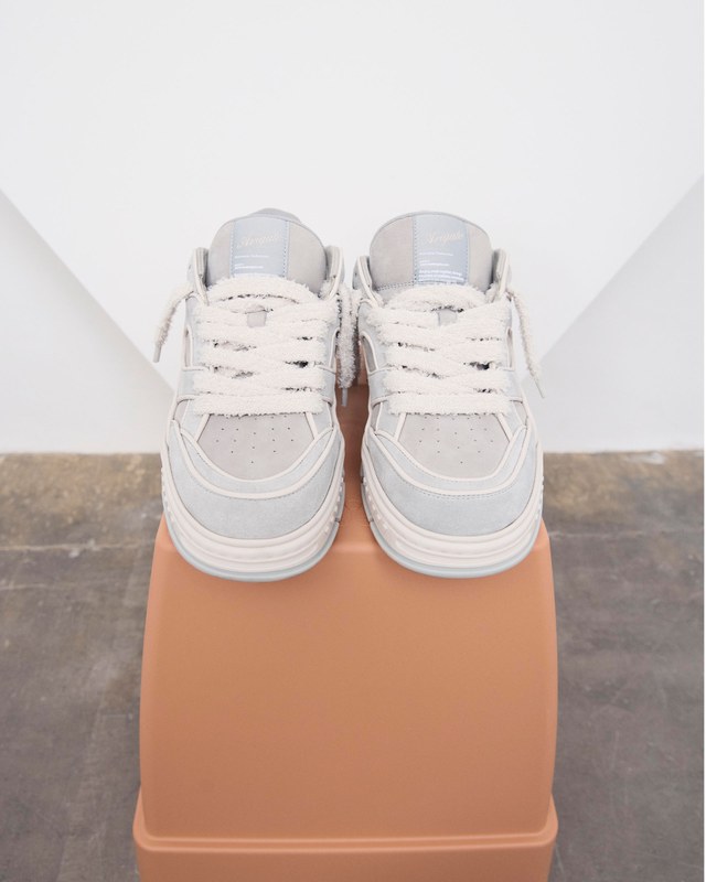 A pair of white sneakers with thick laces displayed on a rectangular, brown platform against a neutral background.