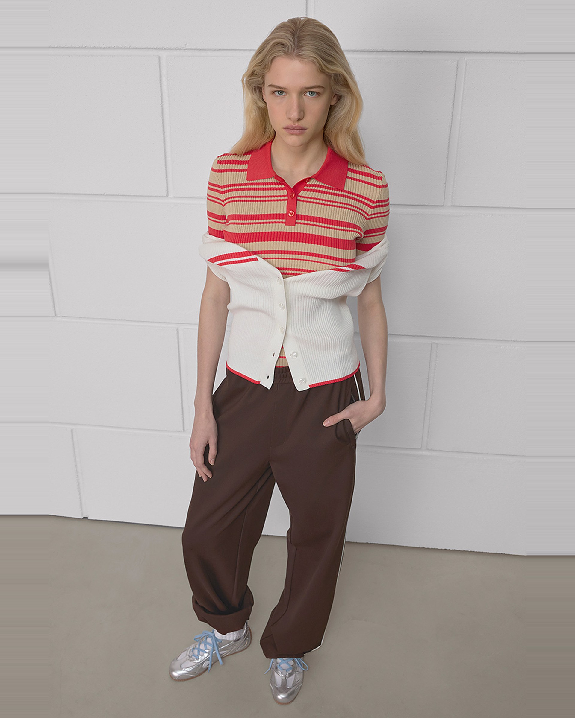 A person wearing a red striped polo, white sweater, brown pants, and silver shoes stands against a white brick wall, looking at the camera.