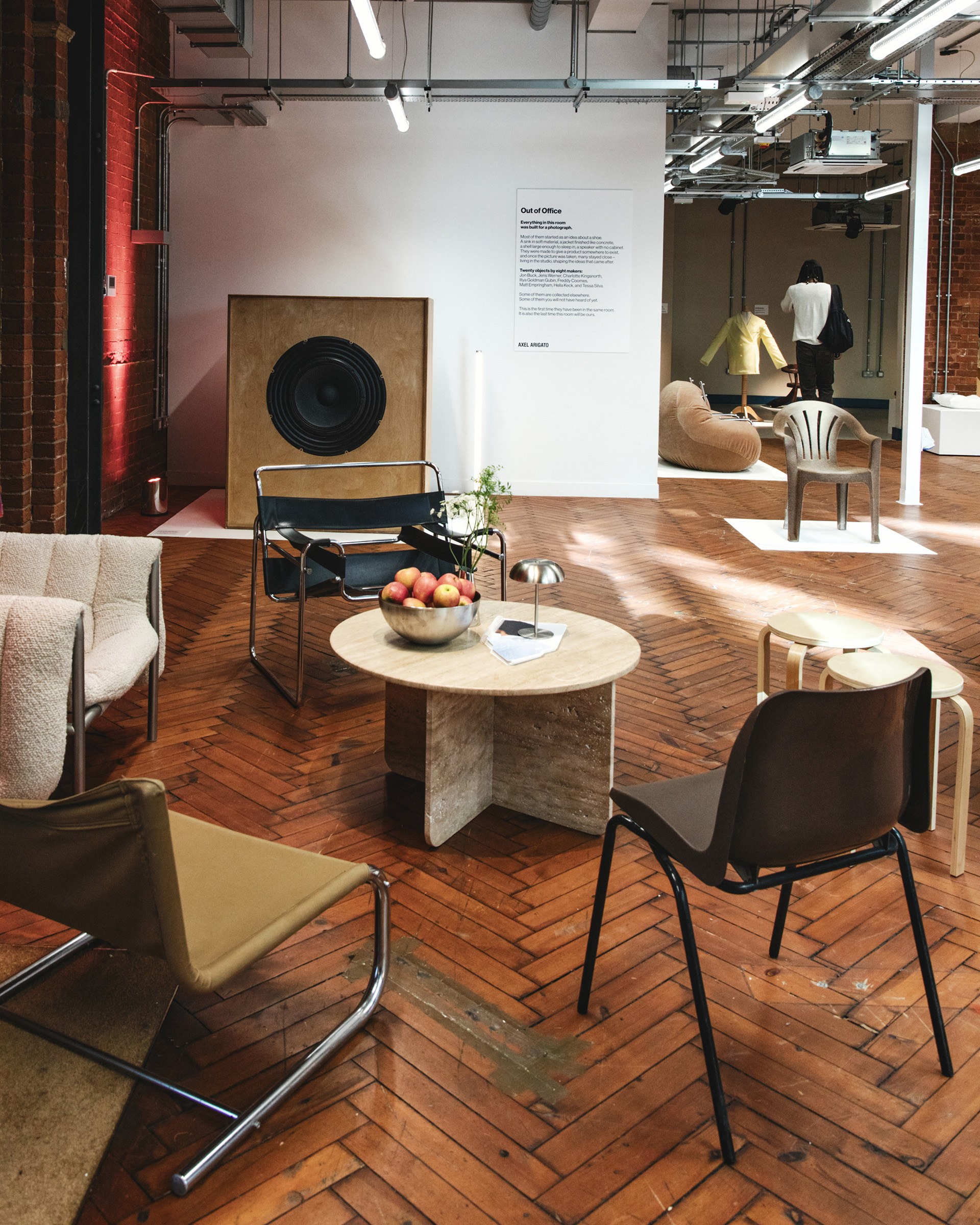 Modern lounge area with assorted chairs, a round wooden table holding a fruit bowl, and abstract art pieces under industrial lighting.