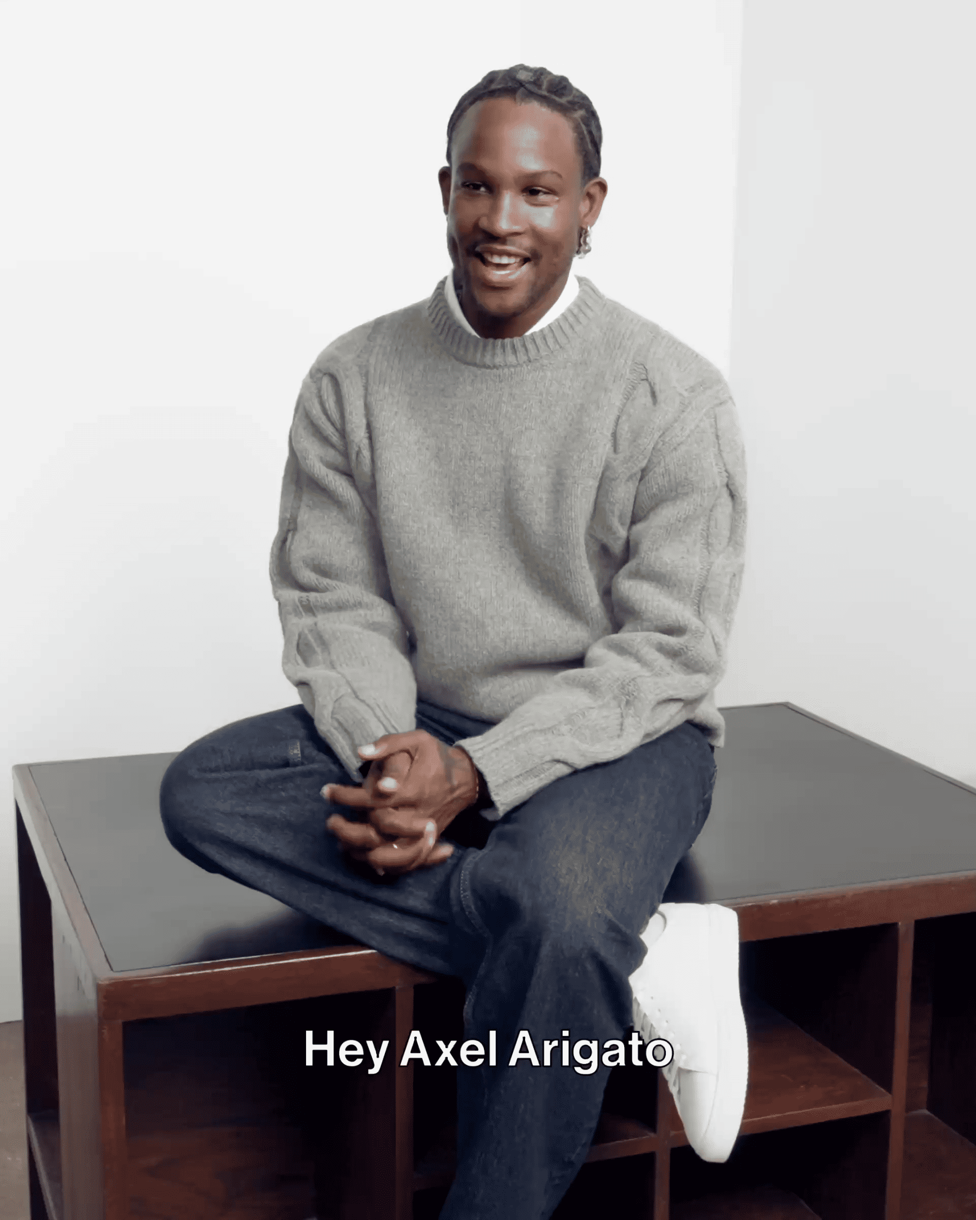 A person wearing a gray sweater and jeans sits on a wooden table, smiling and clasping hands, with the text "Hey Axel Arigato" below.