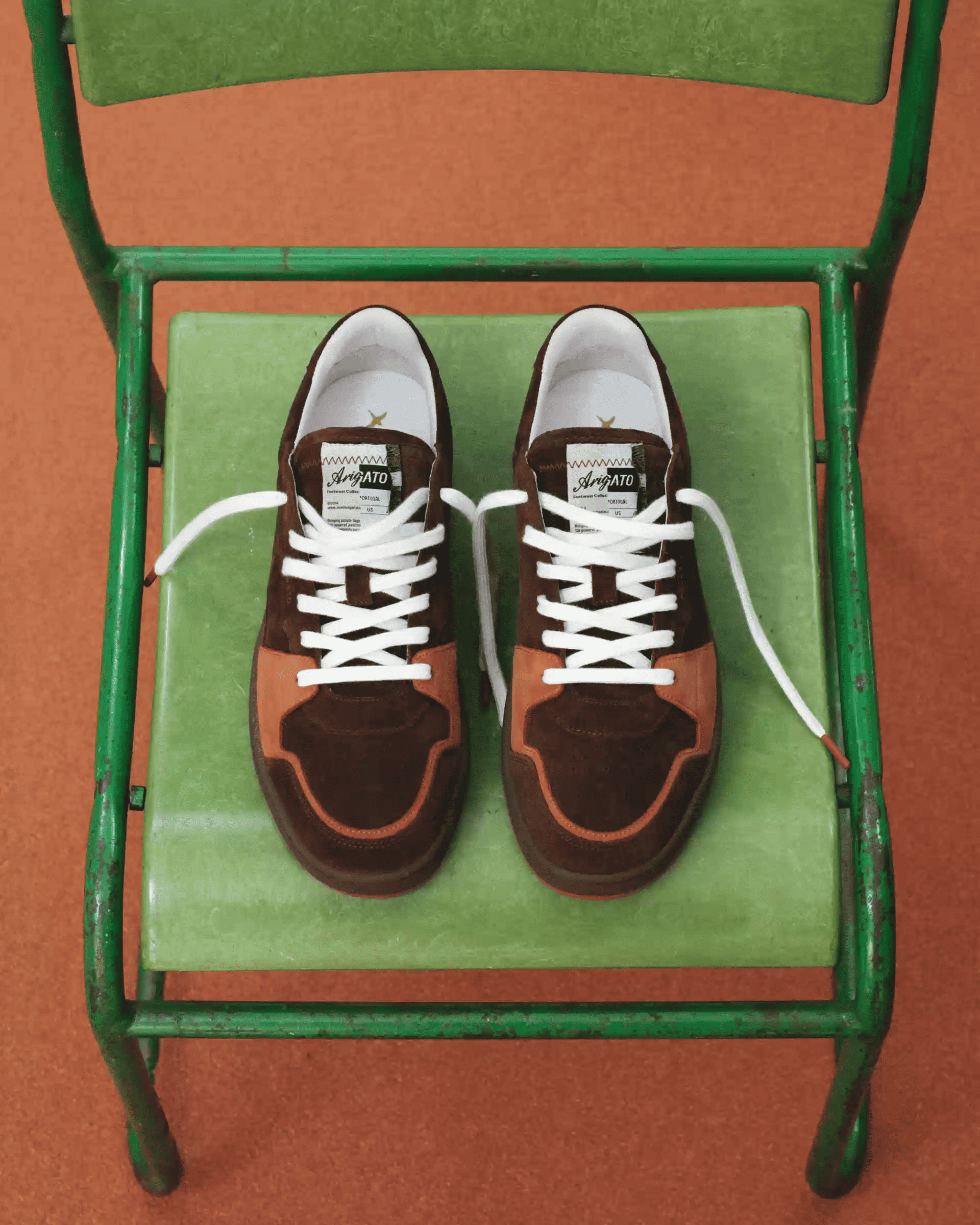 Brown and orange sneakers with white laces placed on a green chair against an orange background, viewed from above.
