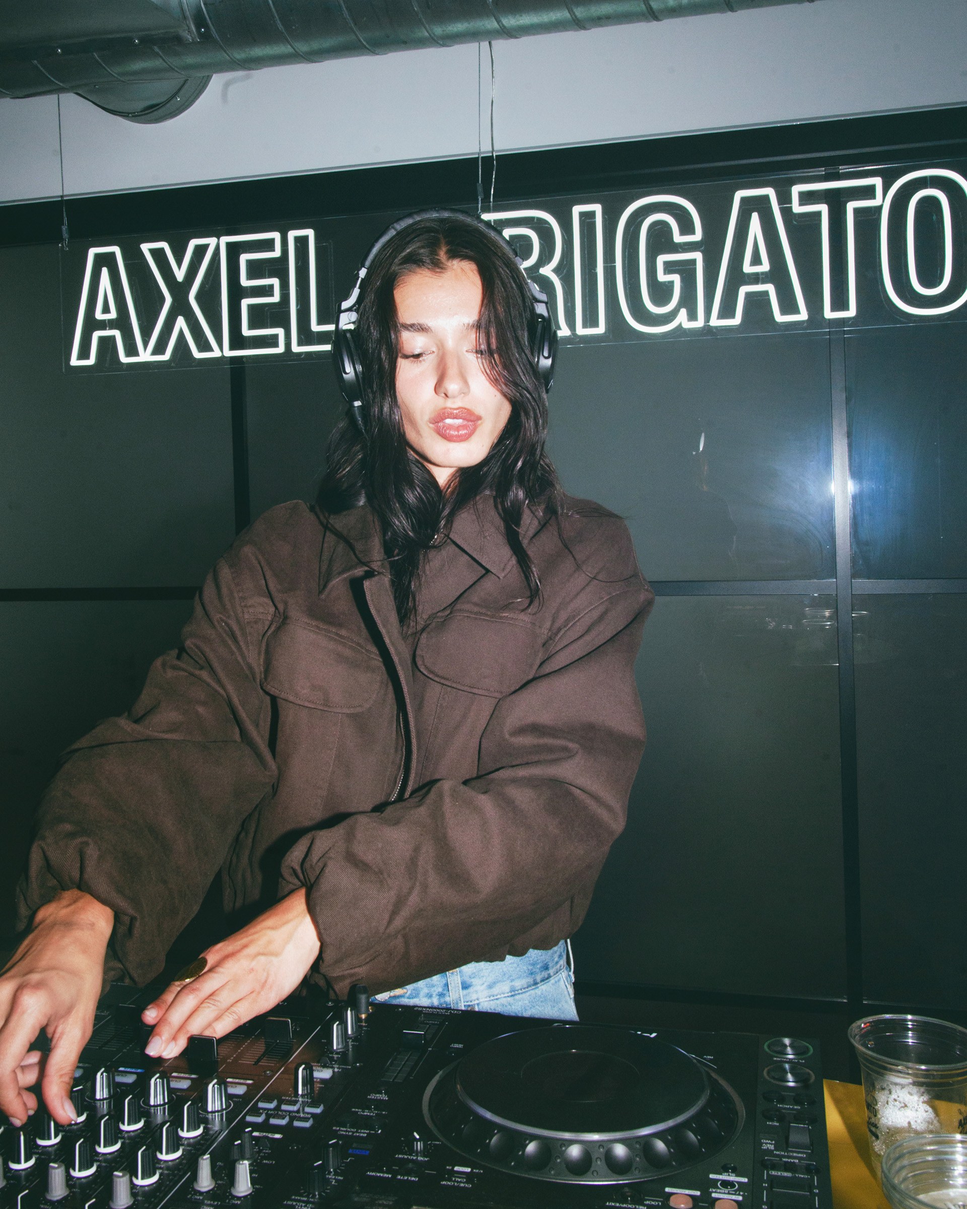 A DJ with headphones stands behind a mixing console, adjusting controls. A sign reading "Axel Arigato" is visible in the background.