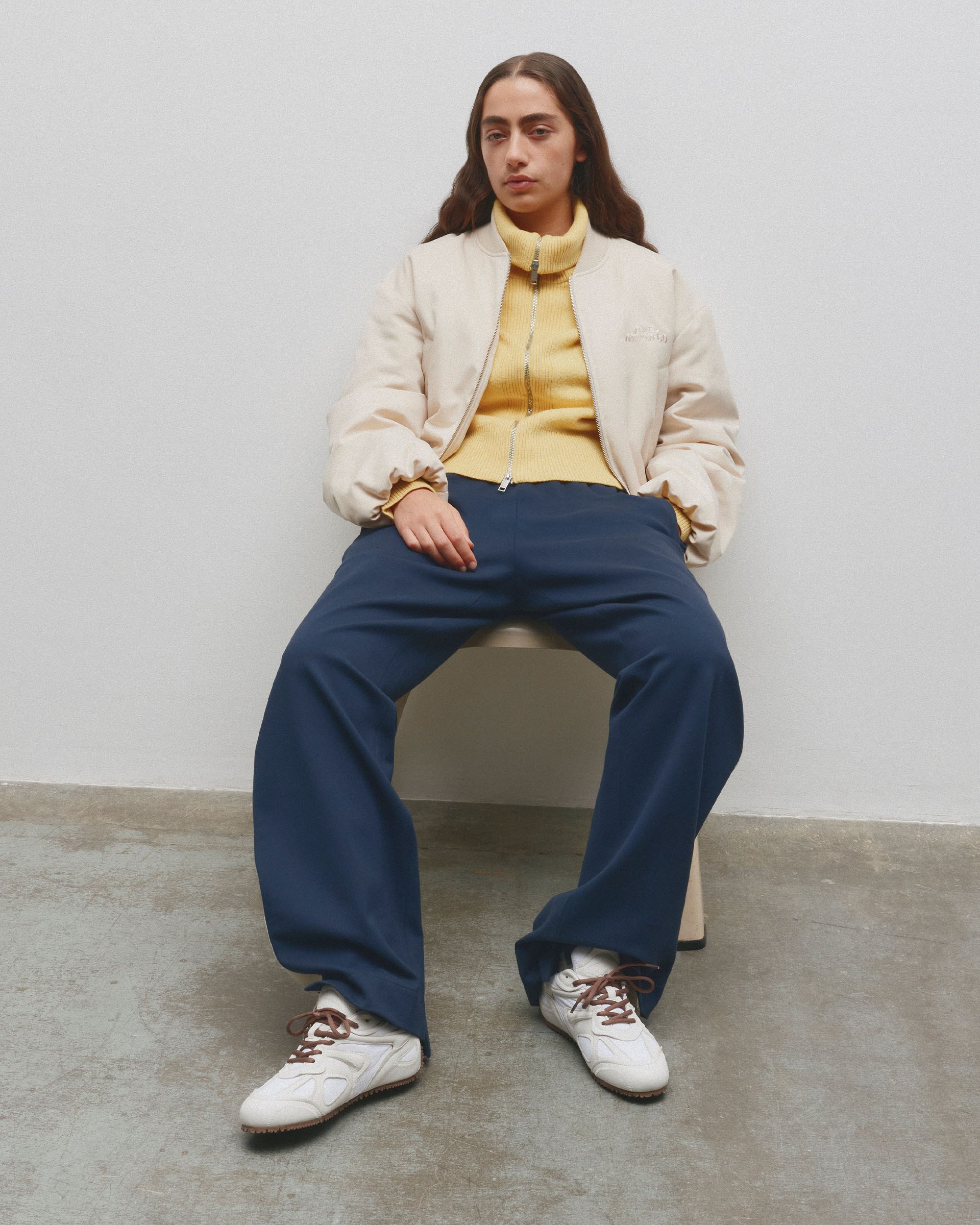 Person sitting on a bench, wearing a beige jacket, yellow sweater, navy pants, and white sneakers, against a plain white wall.