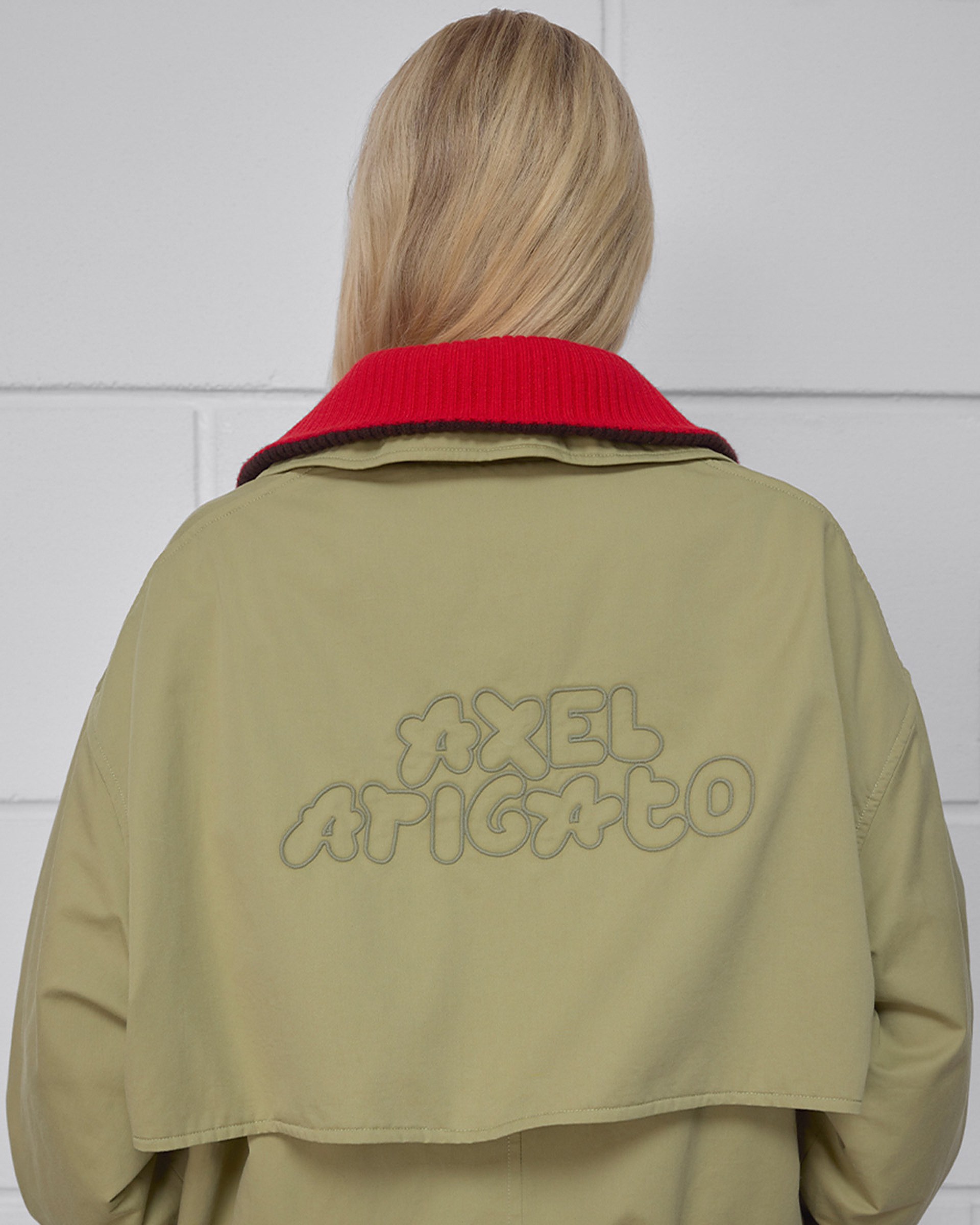 Person with long blonde hair wearing a beige coat with "Axel Arigato" embroidered on the back in stylized text, featuring a red collar.