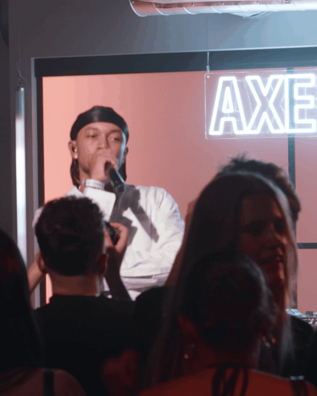 A musician wearing a durag and white shirt performs on stage with a microphone, in front of a crowd, with "AXE" sign lit in the background.
