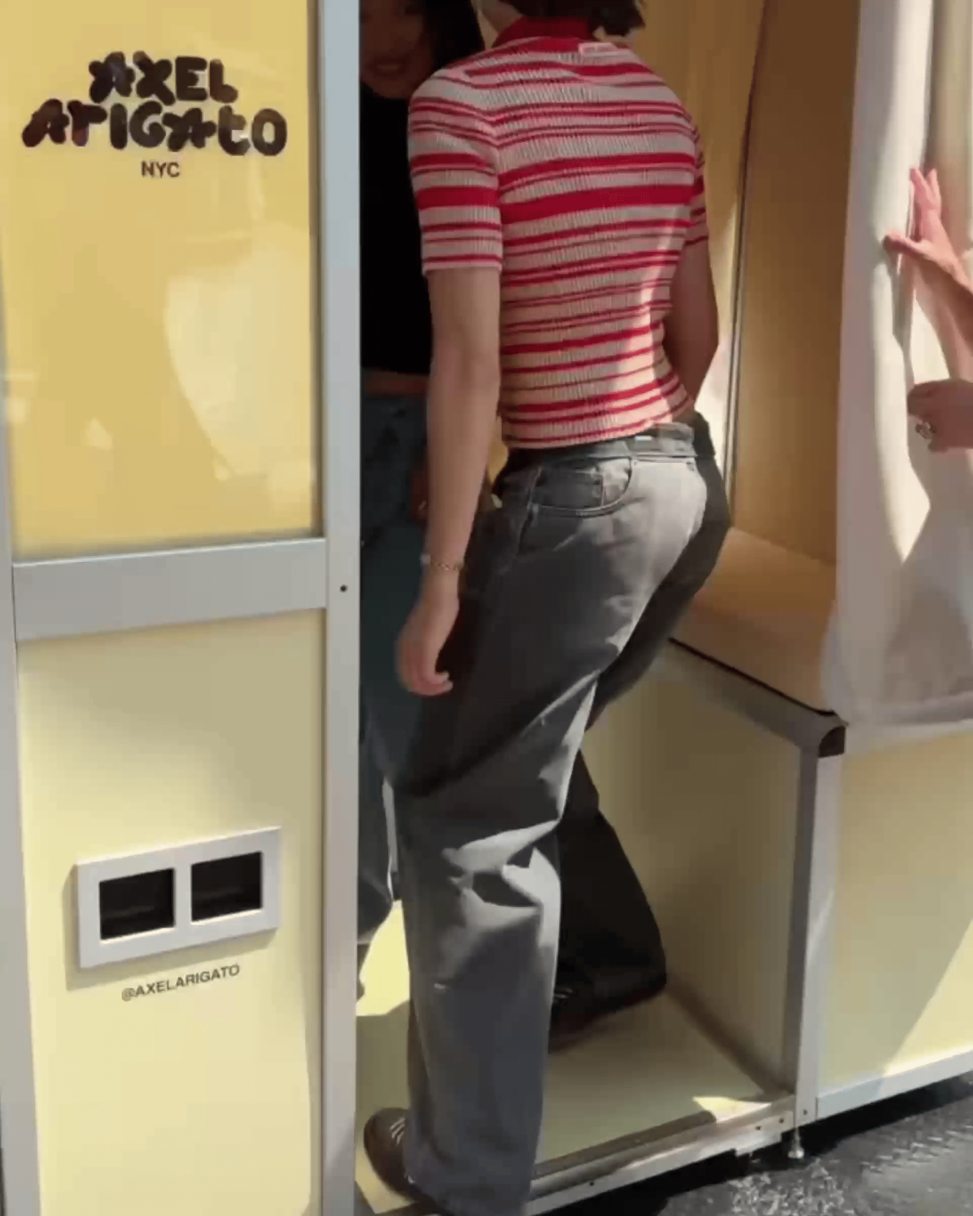 Person in a red striped shirt and jeans stepping inside a yellow booth with "Axel Arigato" branding, while another person holds the curtain.