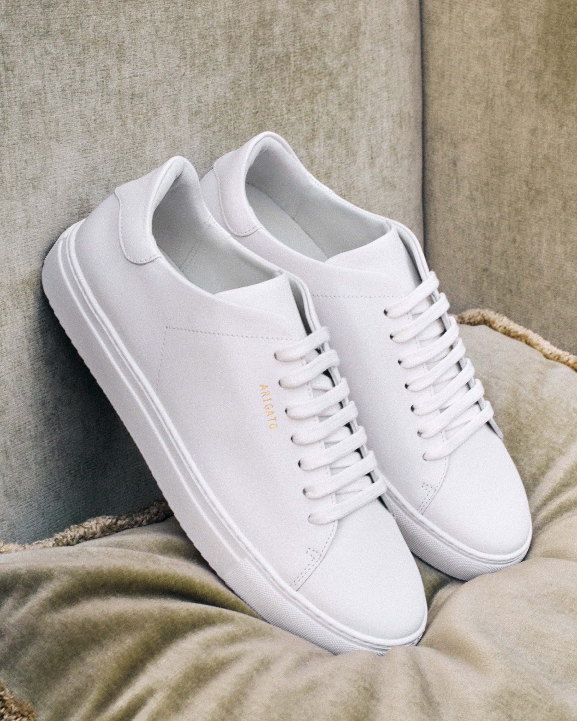 White sneakers with clean design, gold lettering on the side, and white laces, placed on a beige cushioned surface.