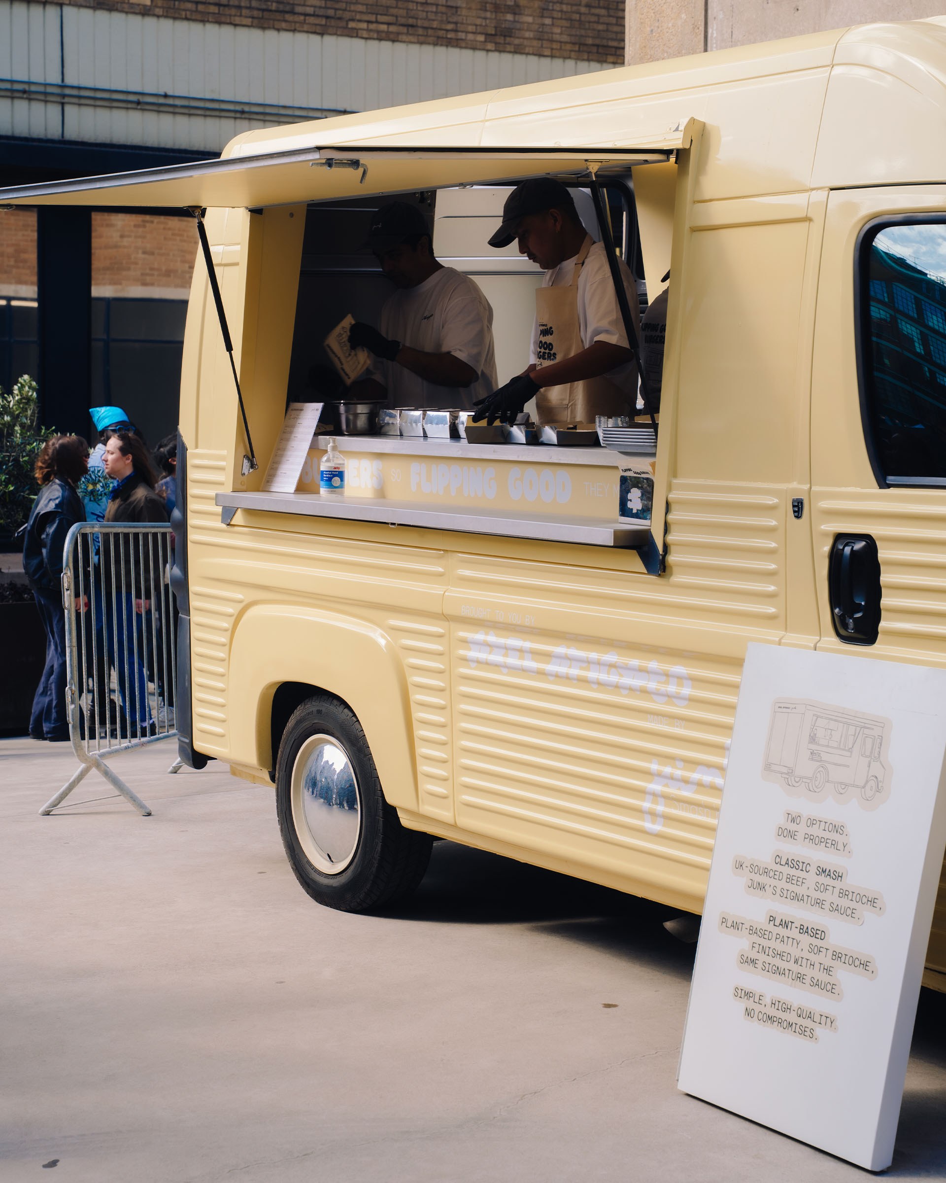 Yellow food truck with open serving window. Two people inside preparing food. Menu board stands outside. Crowds gather in the background.