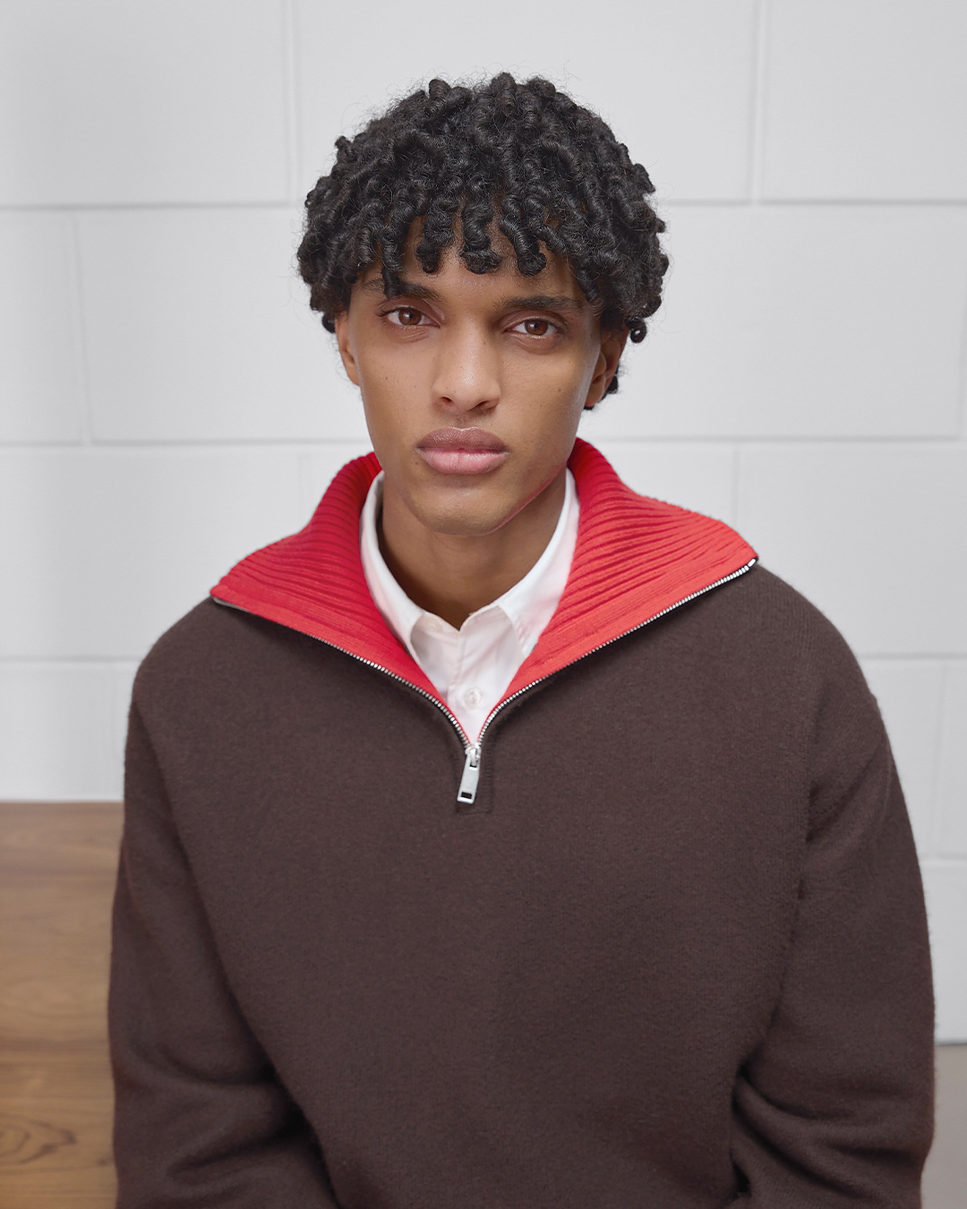 Person with curly hair wearing a brown sweater with a red collar and a white shirt, sitting against a white brick wall.