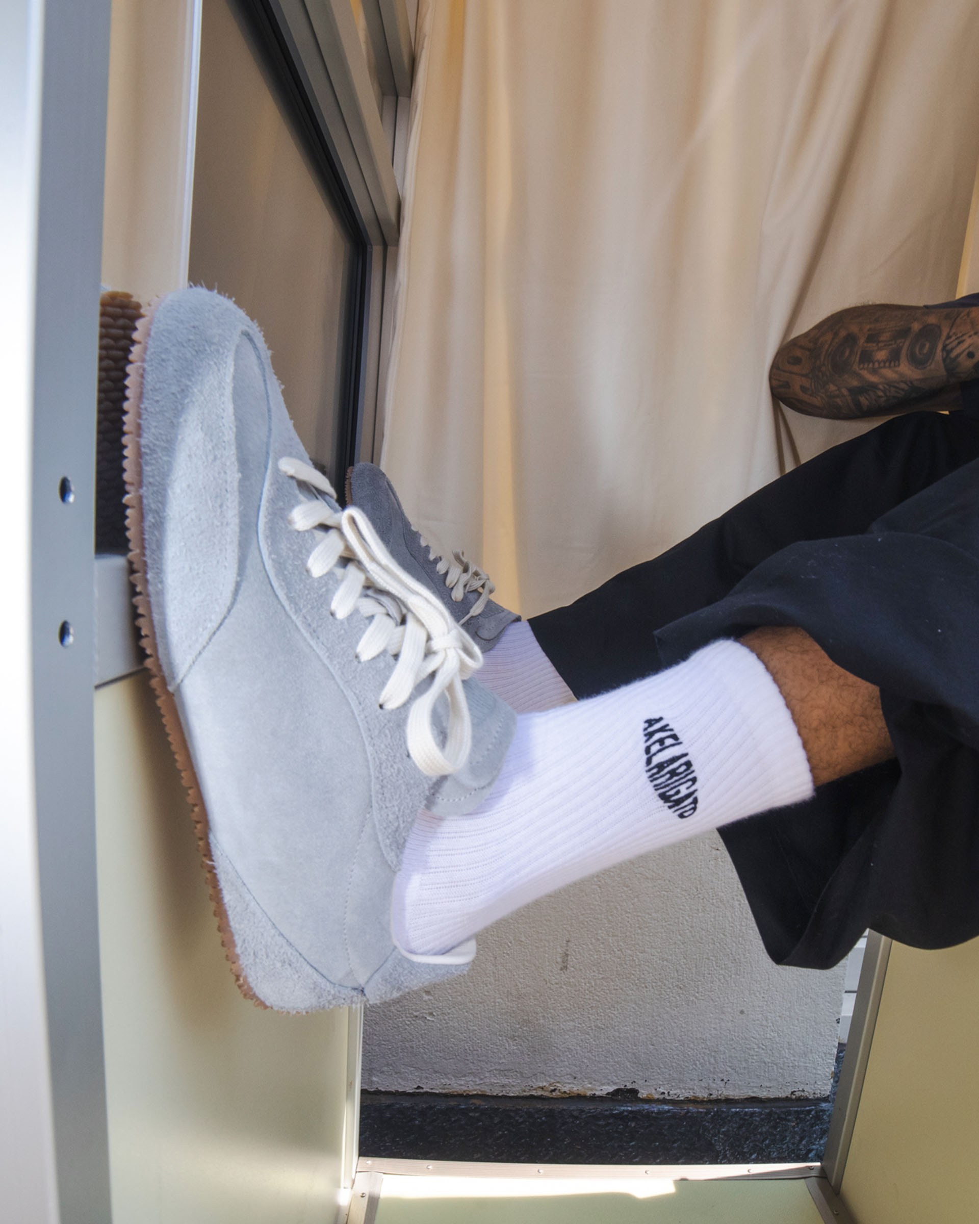Person wearing light gray sneakers and white socks with black text, resting feet on a window ledge. Black pants partially visible.