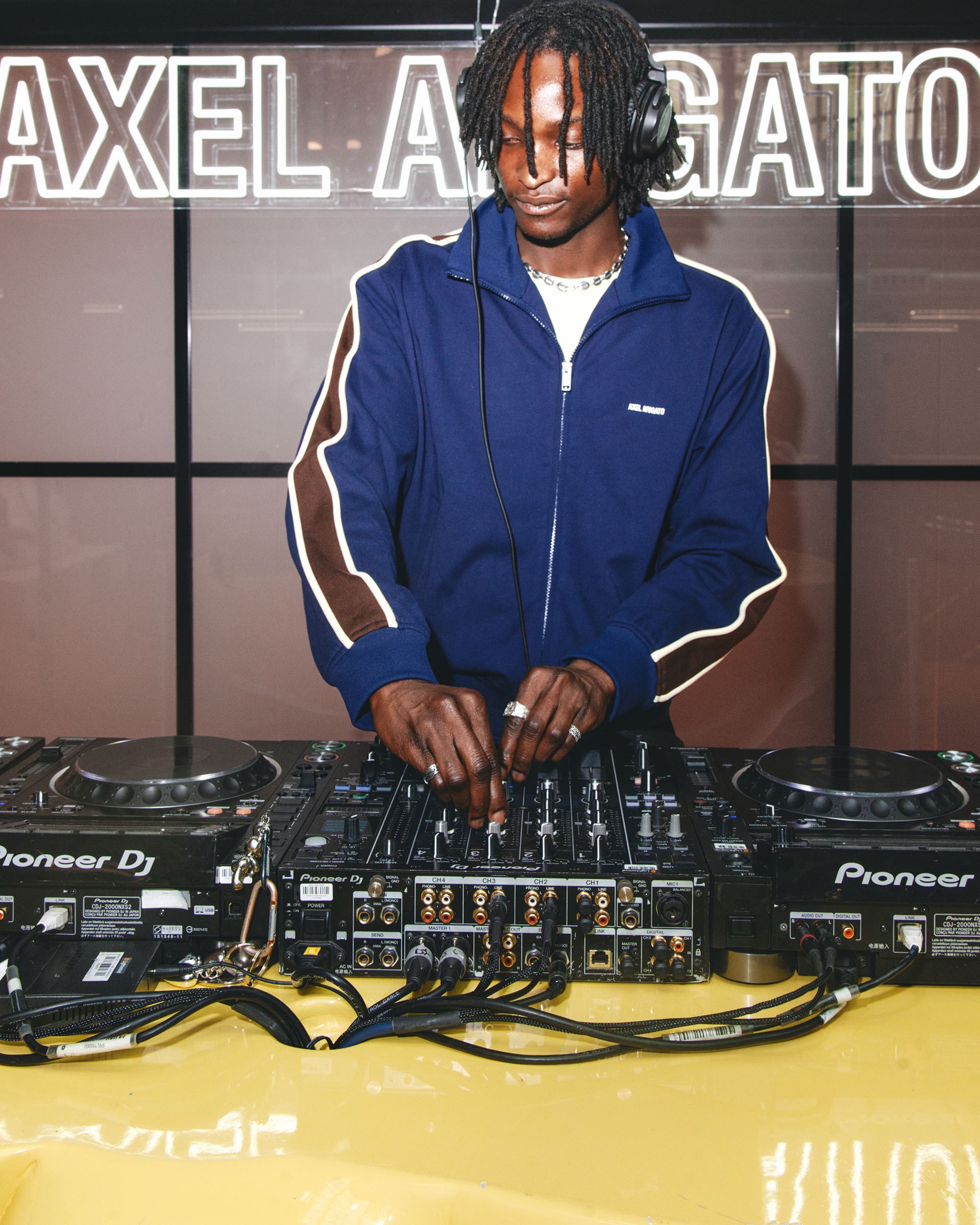 DJ wearing headphones and a navy jacket mixes music on a Pioneer DJ setup against a backdrop with illuminated text.