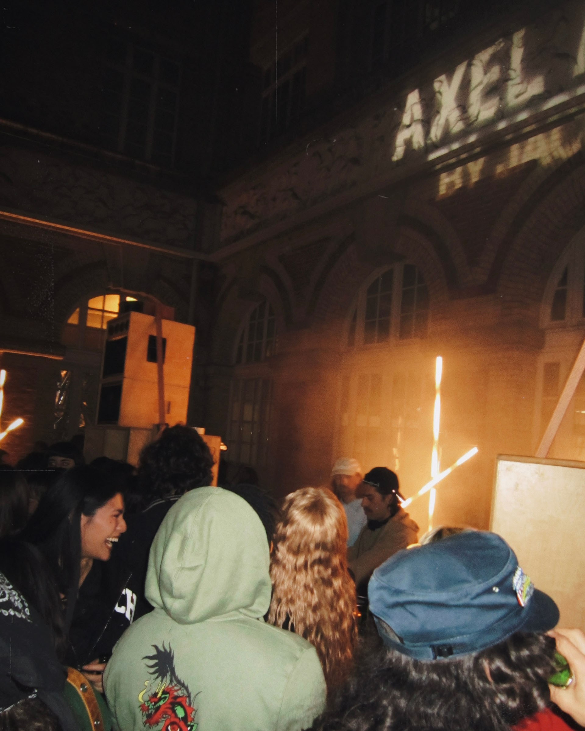 People gathered in a dimly lit courtyard, surrounded by brick walls. Bright lights illuminate the scene, with "AXEL" projected on the building.