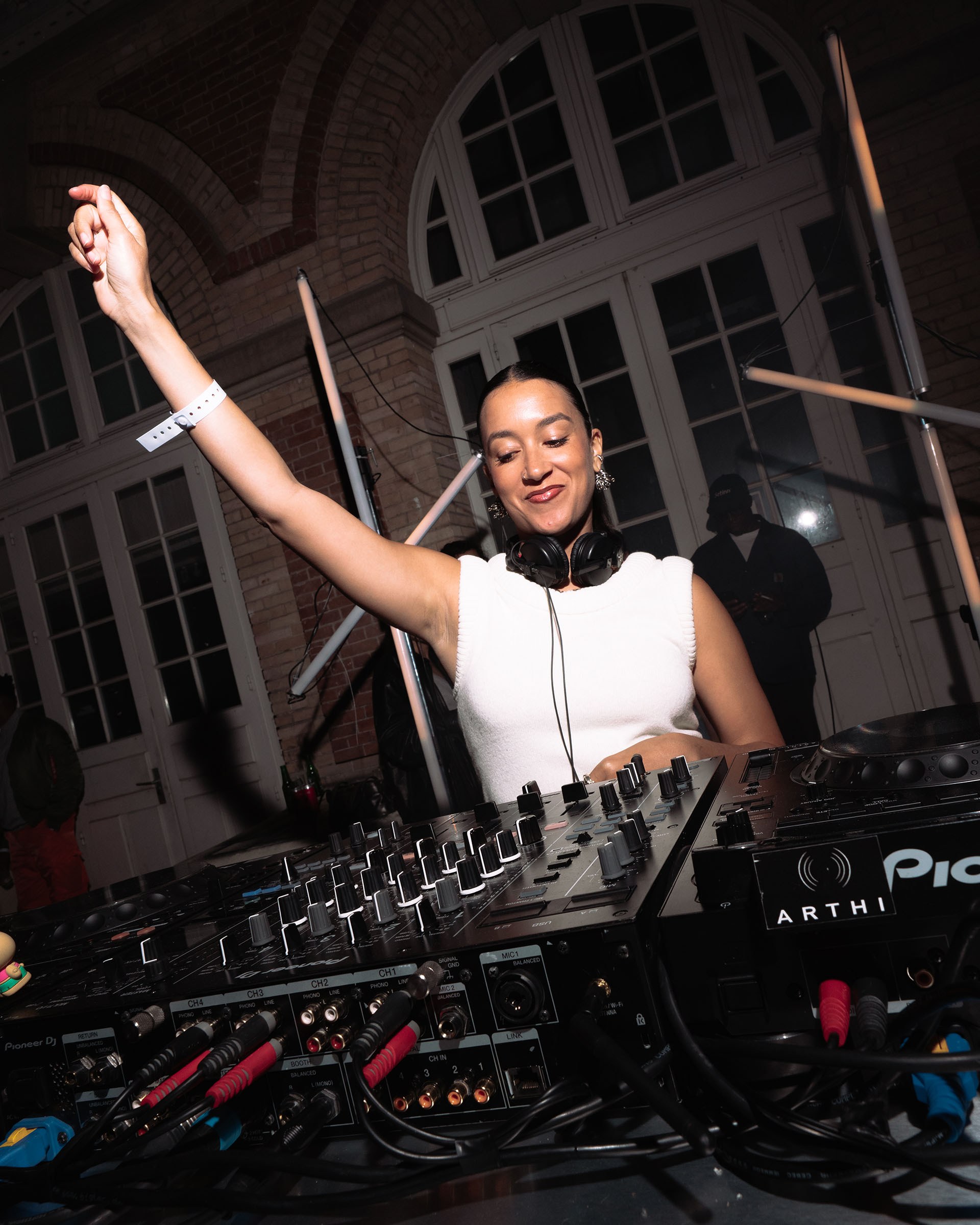 DJ performing energetically with one arm raised at a mixer in a dimly lit venue with arched windows.