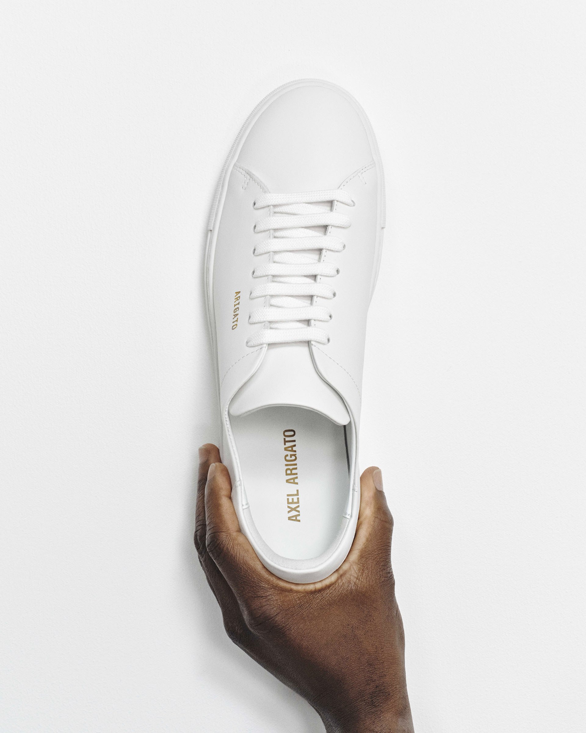 A person holding a white Axel Arigato sneaker with laces, photographed from above, against a plain white background.