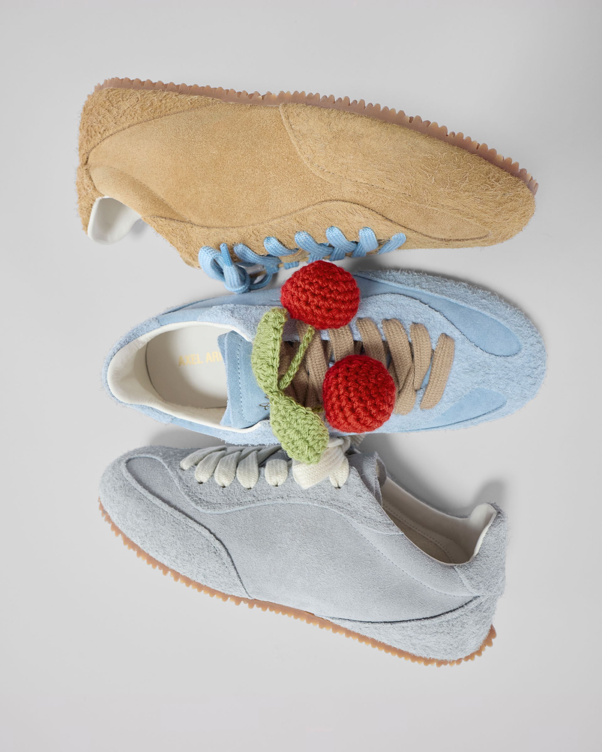 Three suede sneakers: one tan, one light blue with knitted cherries, and one light grey, displayed on a plain background.