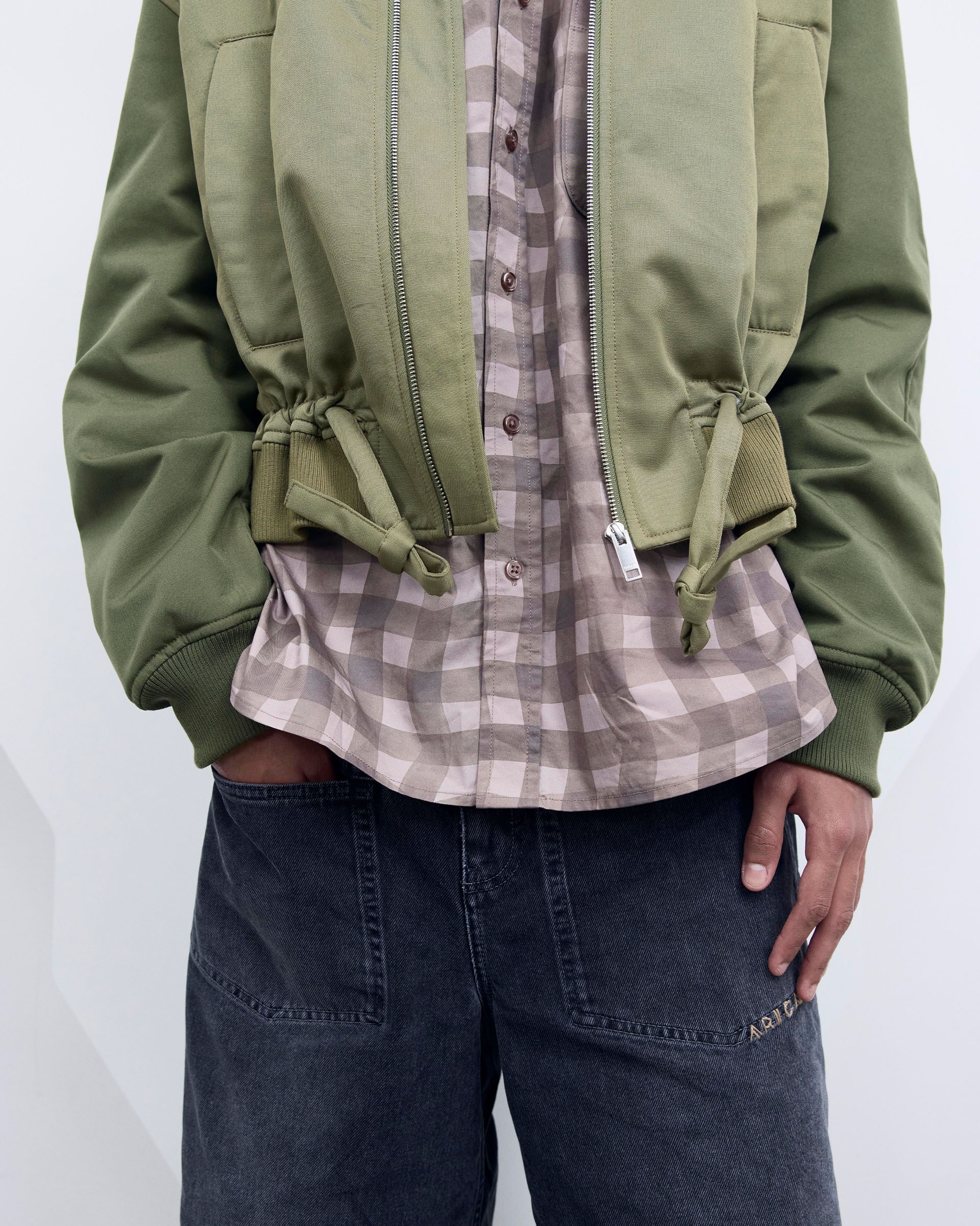 Person wearing a green bomber jacket over a plaid shirt and loose-fitting dark pants, with hands partially in pockets.