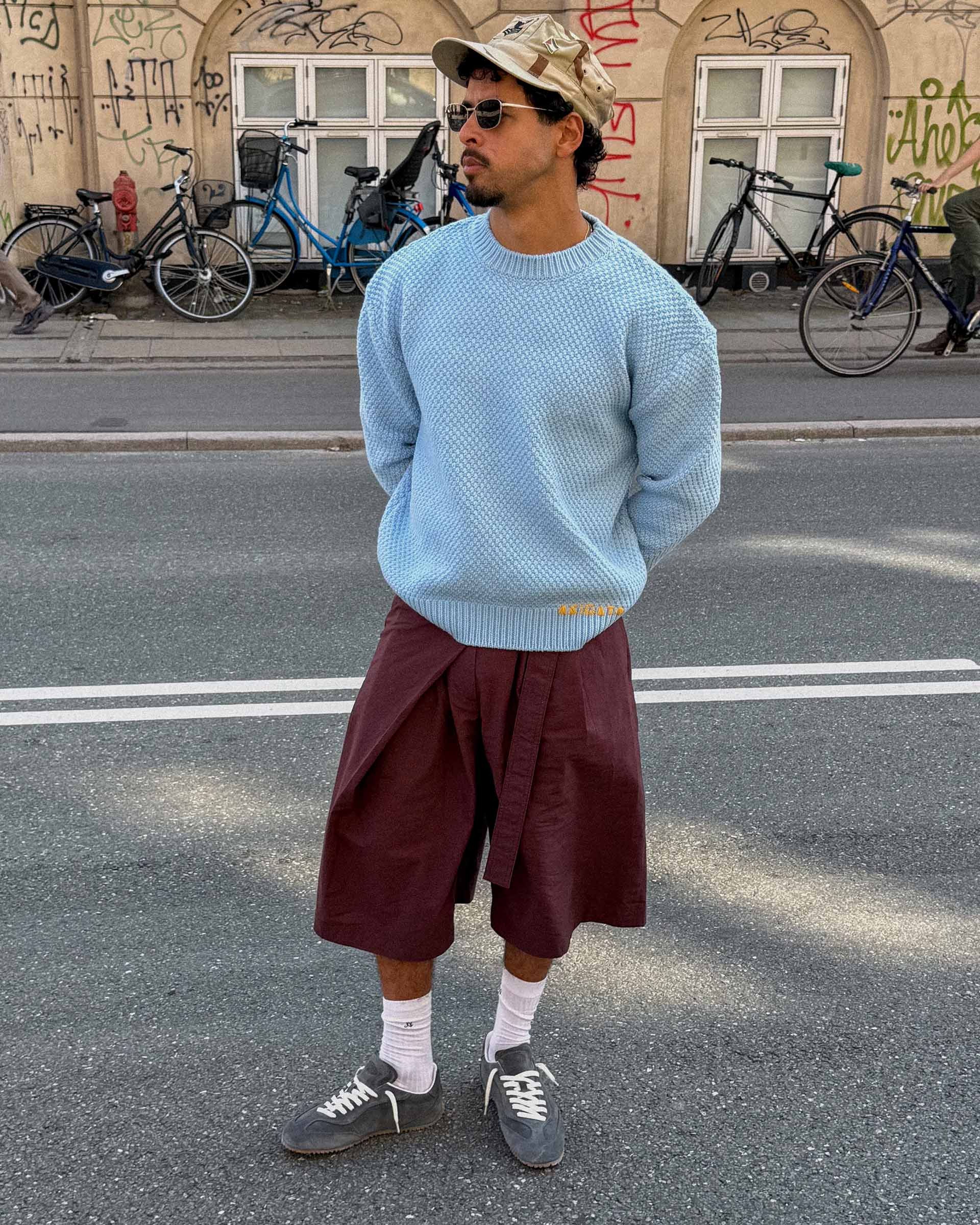Man in a light blue sweater and maroon shorts stands on a street, wearing sunglasses and a cap, with bicycles and graffiti in the background.