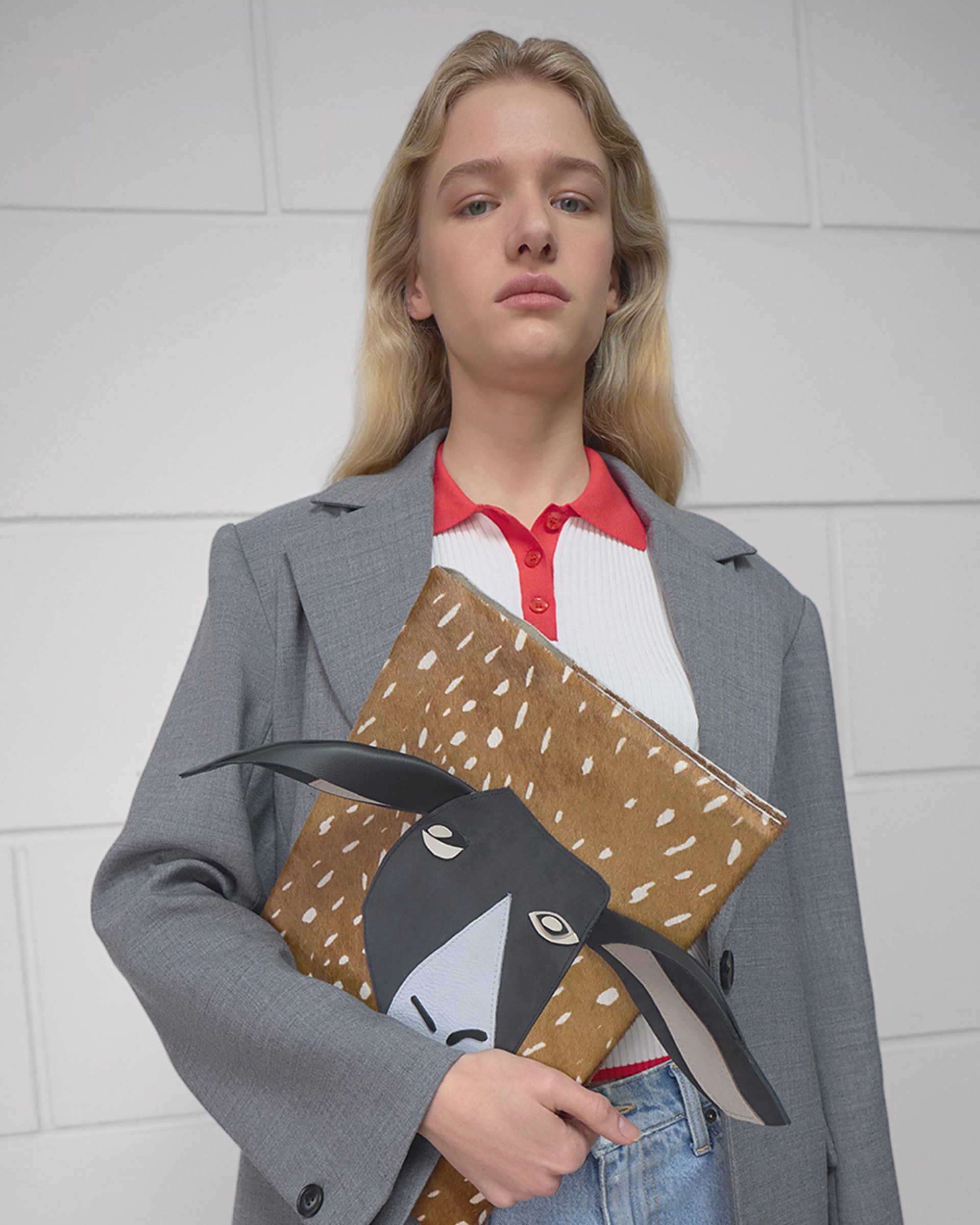 A person in a gray blazer holds a folder with an illustrated goat face on it, standing against a white tiled wall.