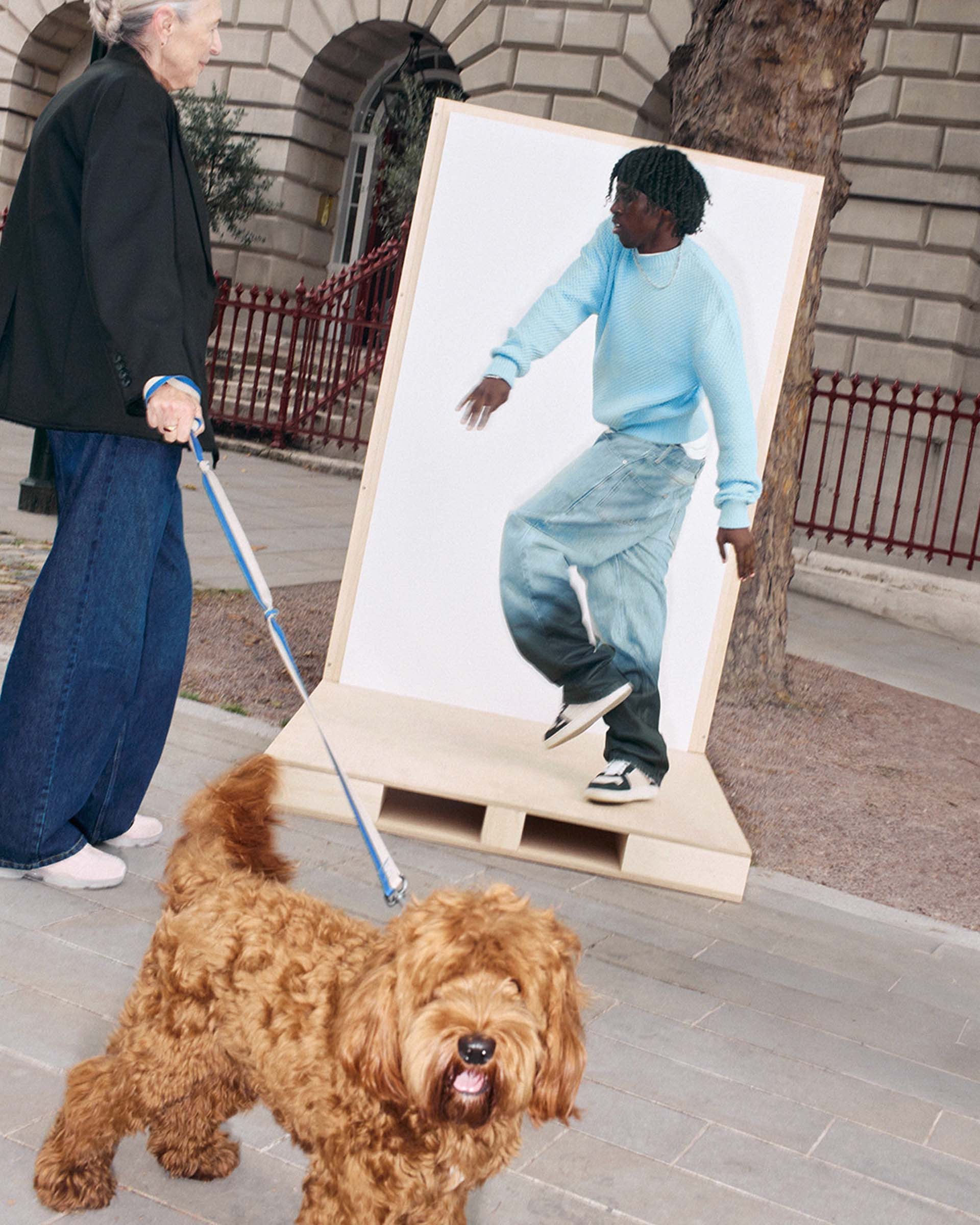 A person walks a curly-haired dog past a large photo of a person in a blue sweater and jeans, displayed on a city sidewalk.