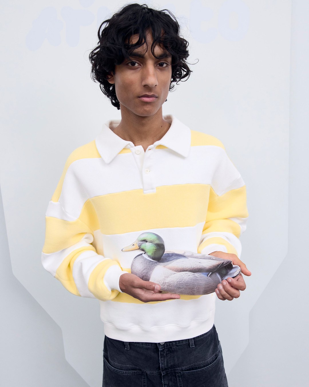 Person with curly hair in a yellow and white striped sweater holding a realistic duck pillow against a plain background.
