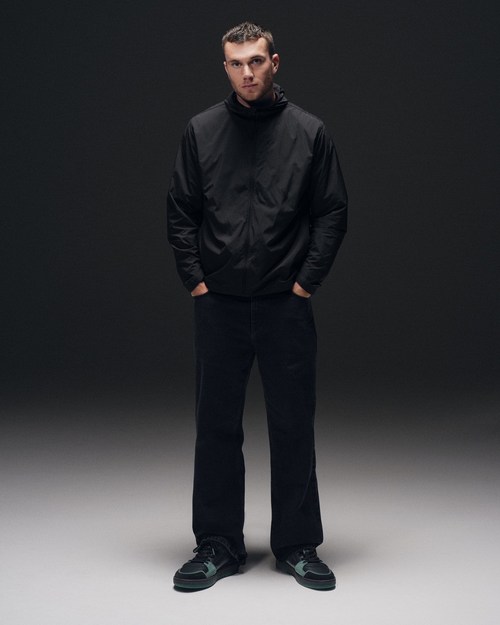 A man stands confidently, wearing a black jacket with a high collar, dark pants, and black sneakers with green accents, set against a dark background.