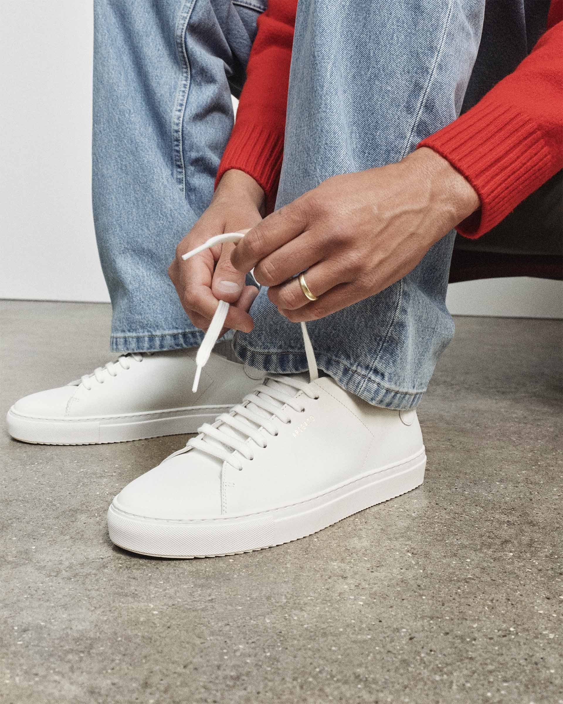 Person in blue jeans and red sweater tying white sneakers on a concrete floor.