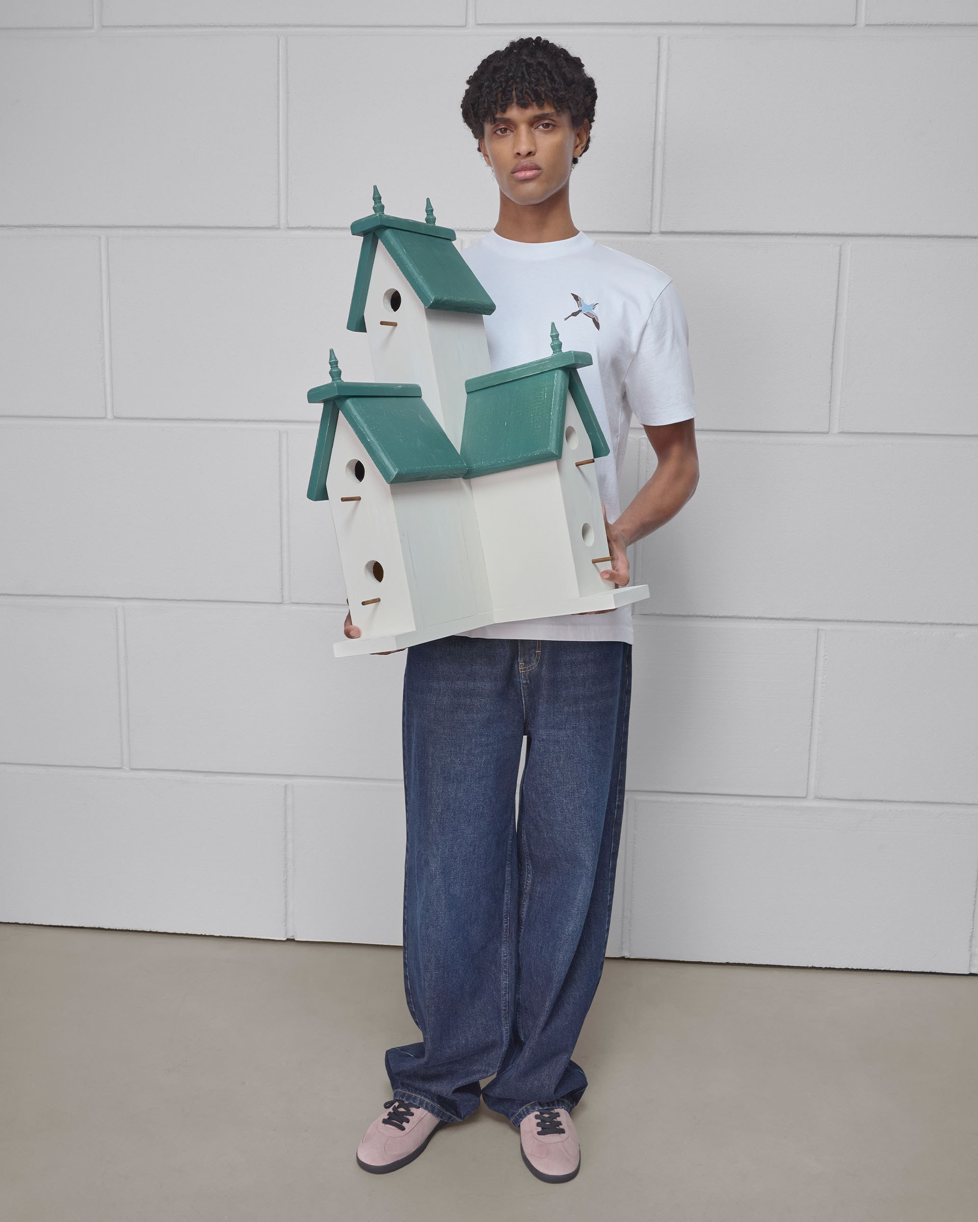 A person in a white T-shirt and jeans holds a large, decorative birdhouse with green roofs, standing against a white brick wall.