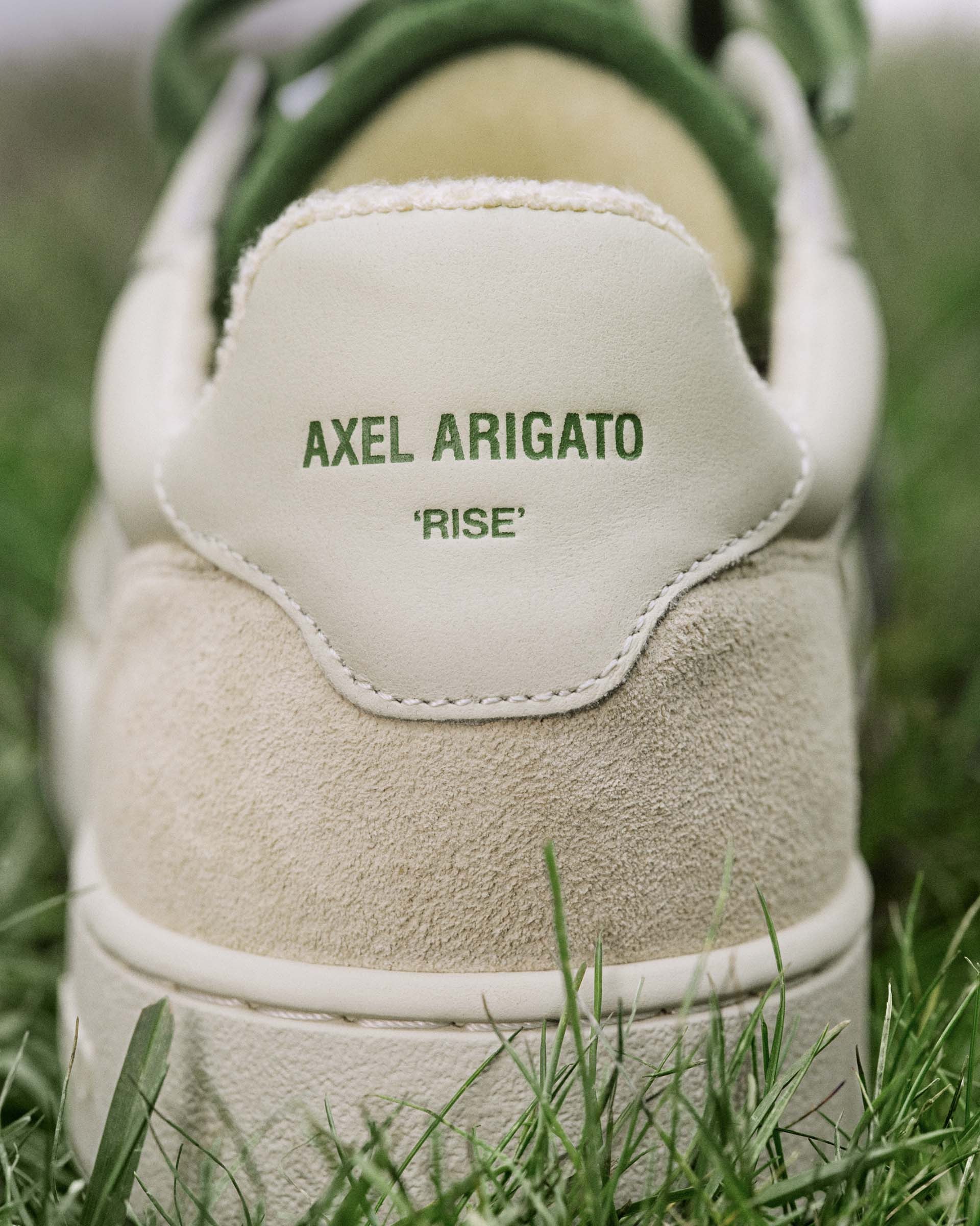 Close-up of an Axel Arigato sneaker heel, featuring cream suede, white leather details, and "RISE" text in green, set on grass.