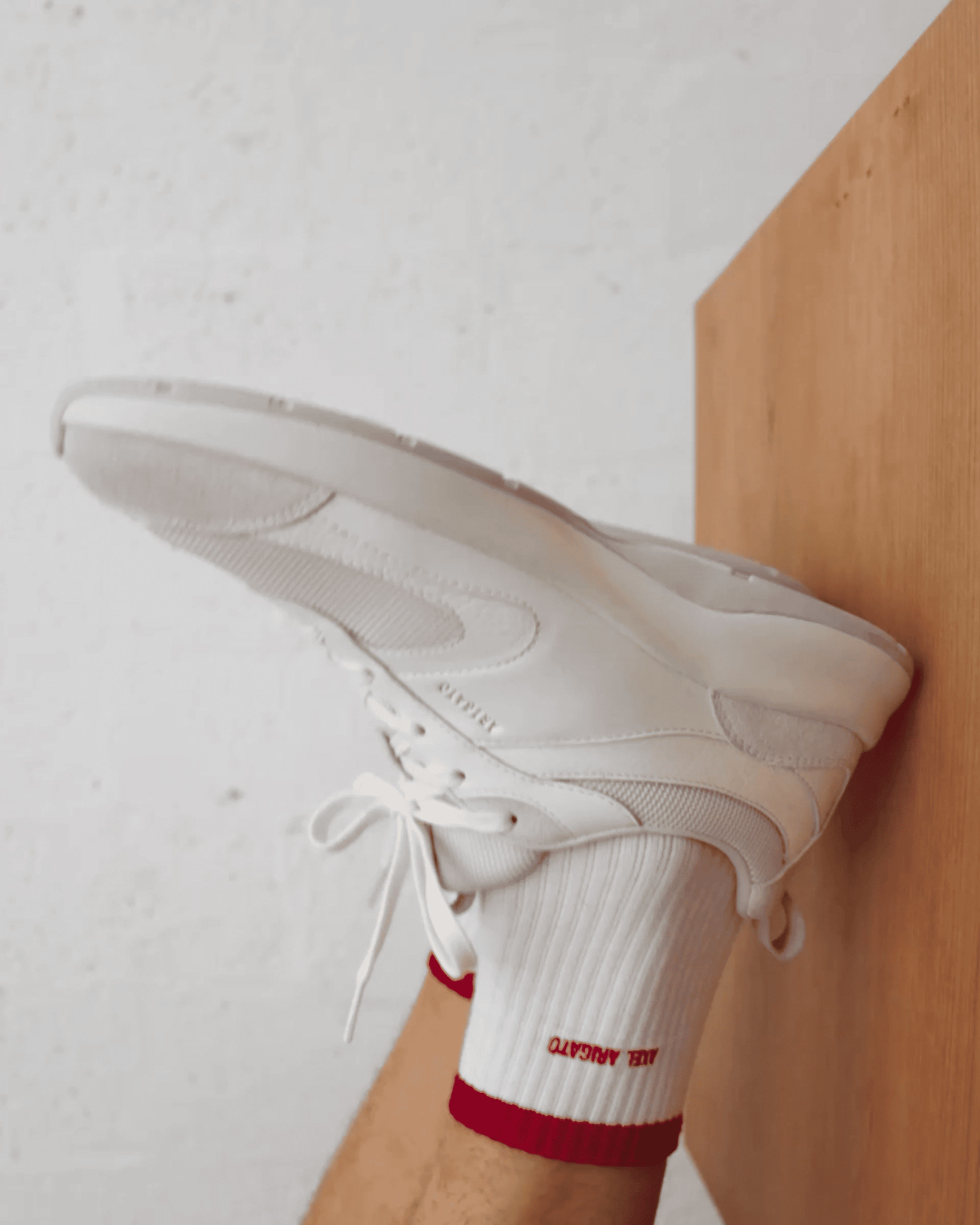 A person wearing white sneakers and white socks with red trim stretches their leg against a wooden surface, with a white wall in the background.
