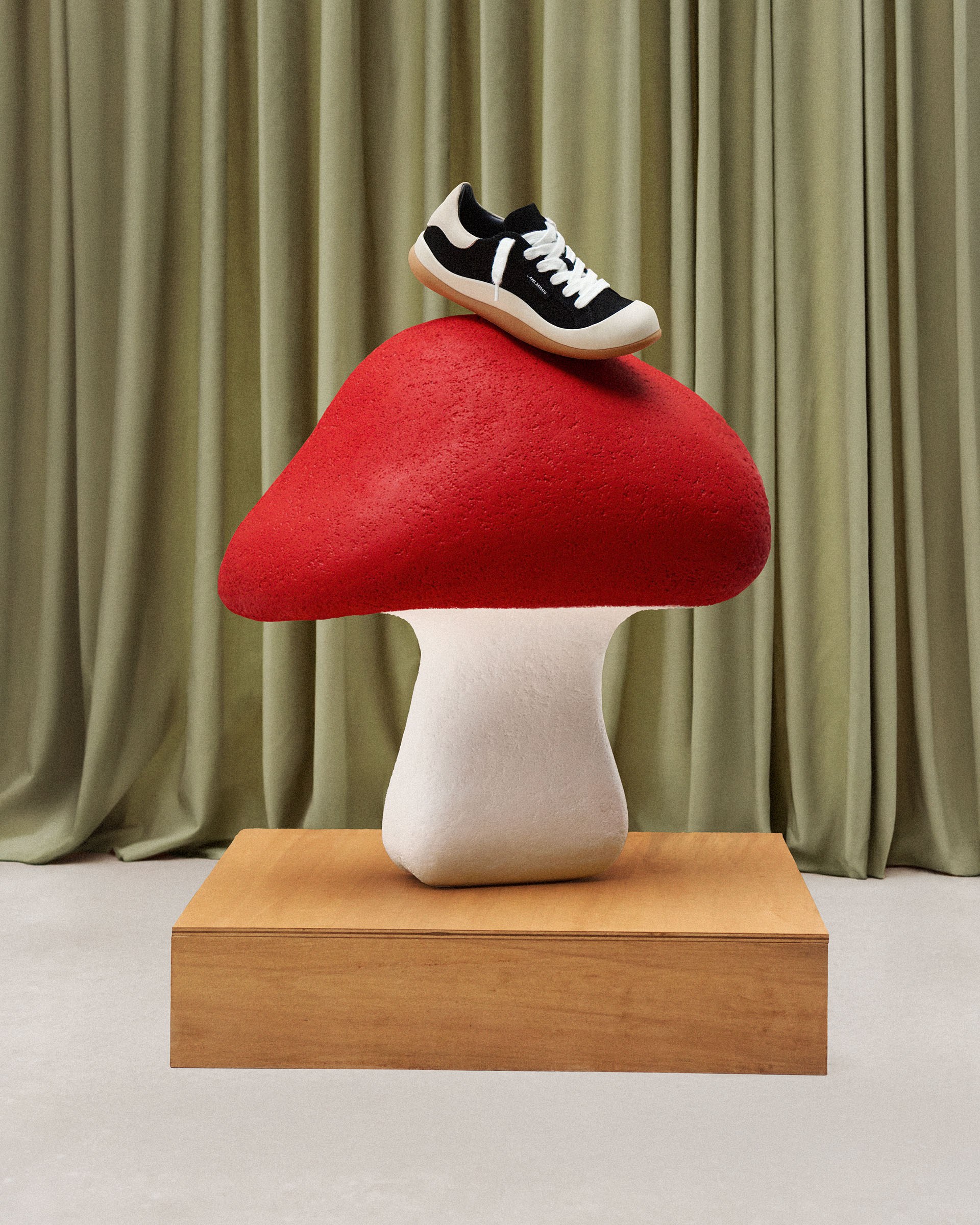 A black and white sneaker displayed on a large red and white mushroom sculpture atop a wooden platform, set against green curtains.