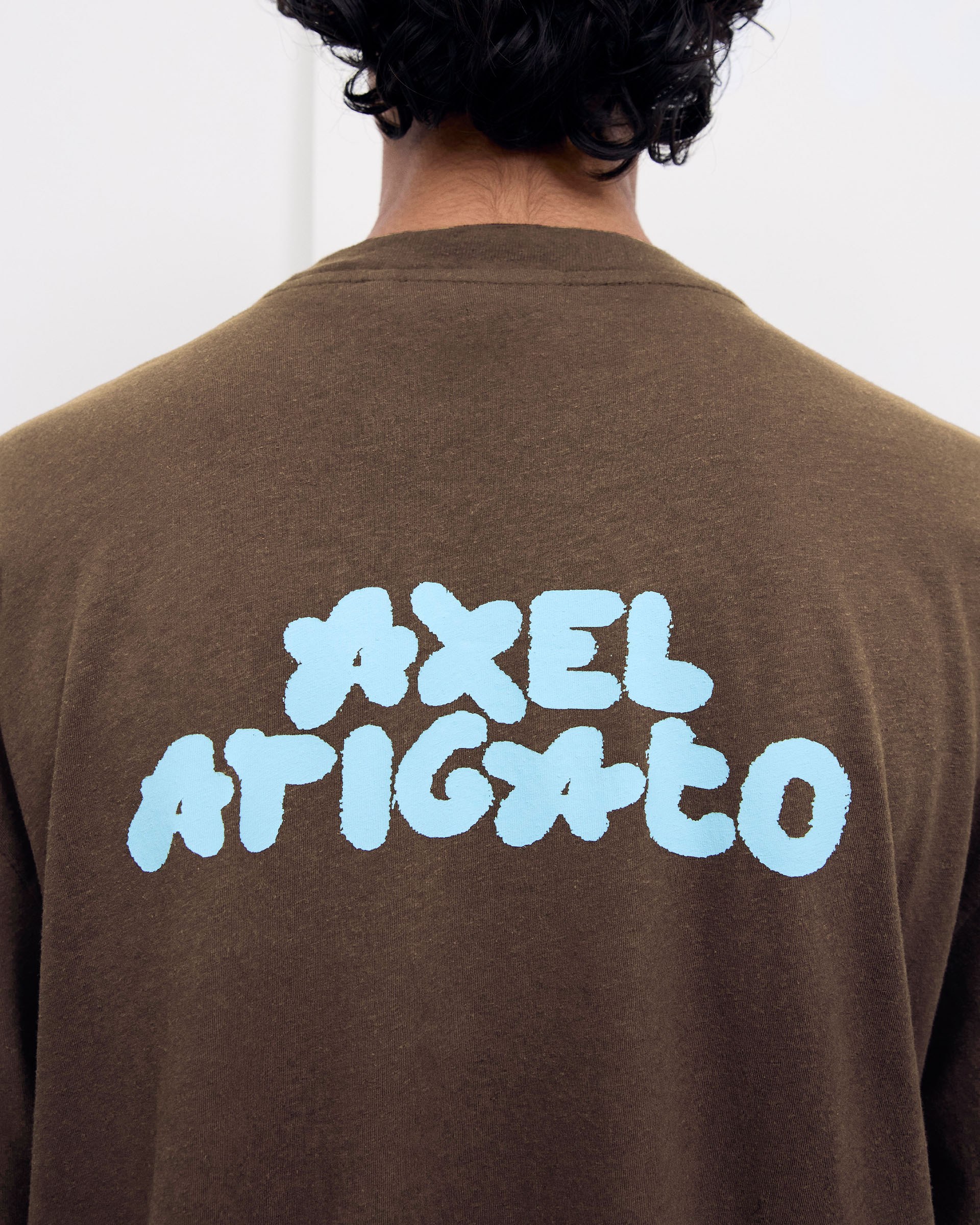 Person wearing a brown shirt with "AXEL ARIGATO" in large, light blue, cloud-like letters on the back.