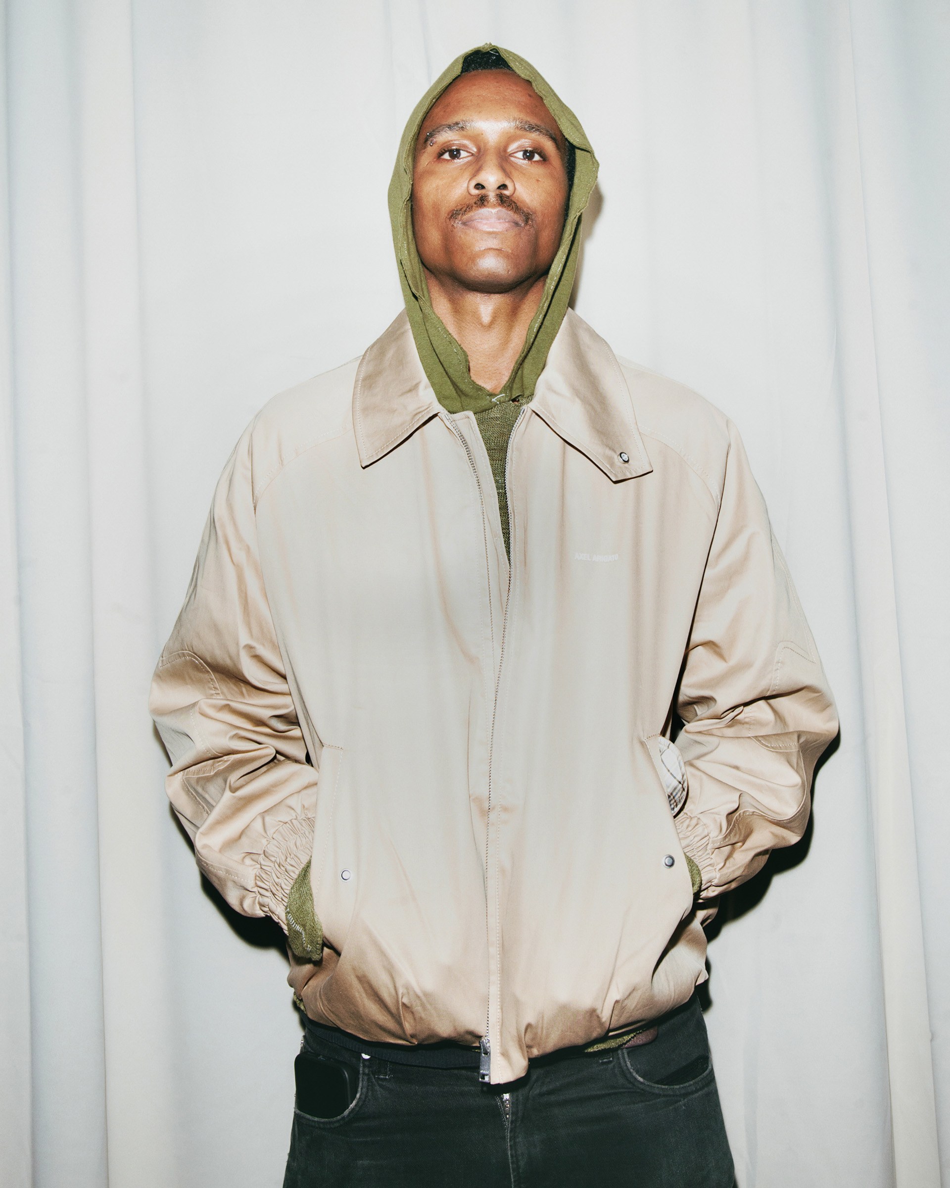 Person wearing a beige jacket over a green hoodie, standing in front of a light-colored curtain, hands in pockets, looking at the camera.