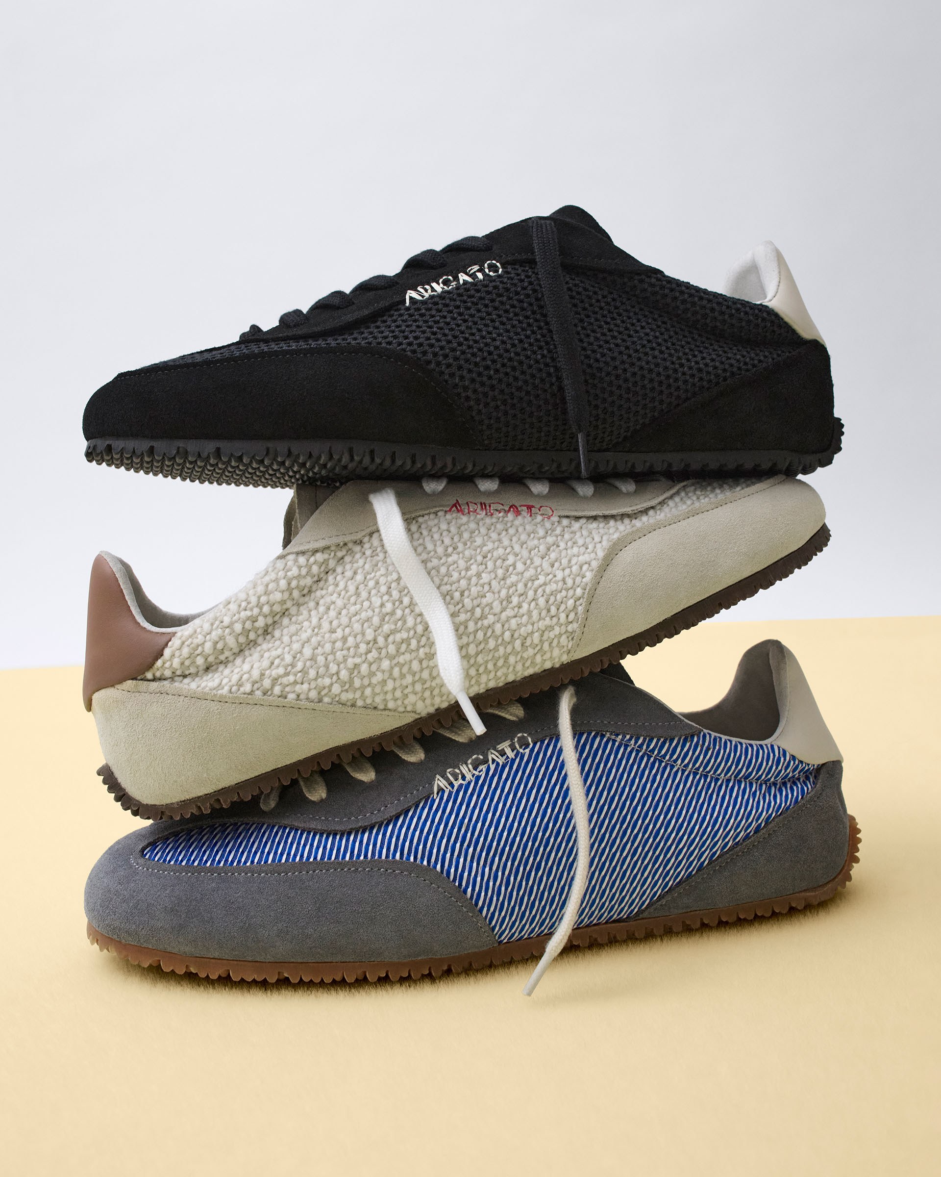 Three stylish sneakers stacked vertically: black mesh, beige with textured fabric, and blue striped, all with rubber soles, on a beige surface.