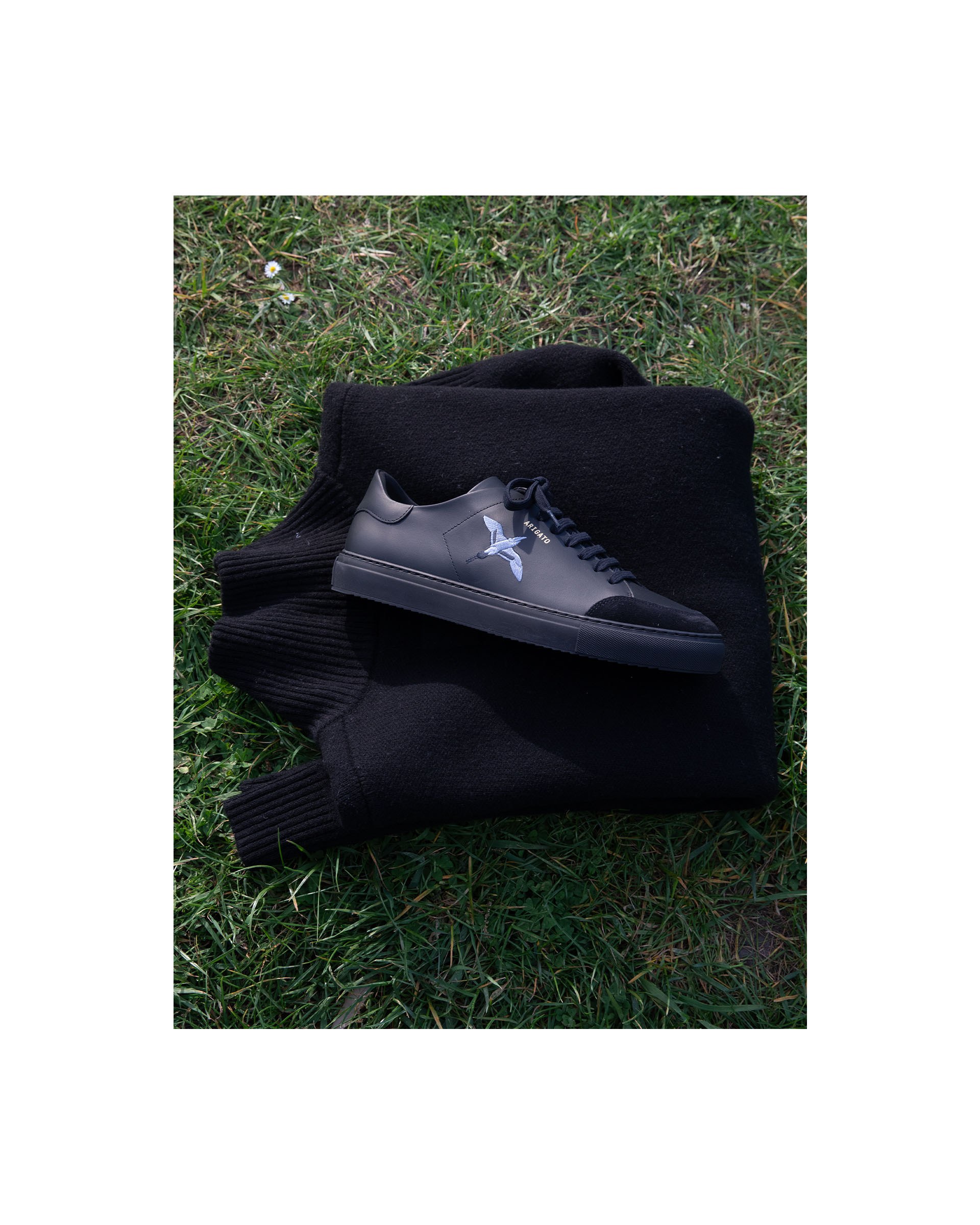 Black sneakers with a white star logo placed on folded black knitwear, lying on the grass.