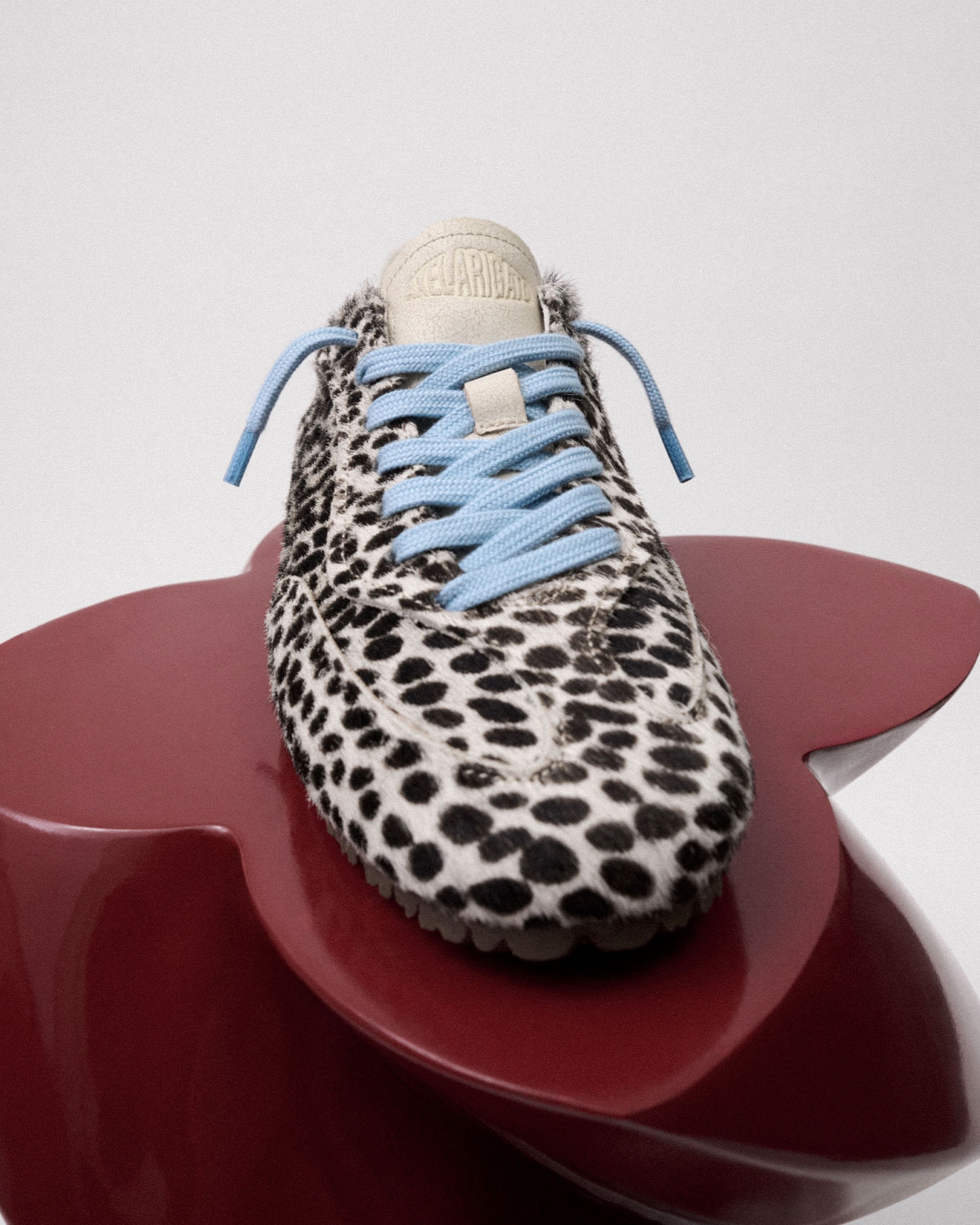 Leopard print sneaker with pale blue laces displayed on a glossy, red, petal-shaped surface against a white background.