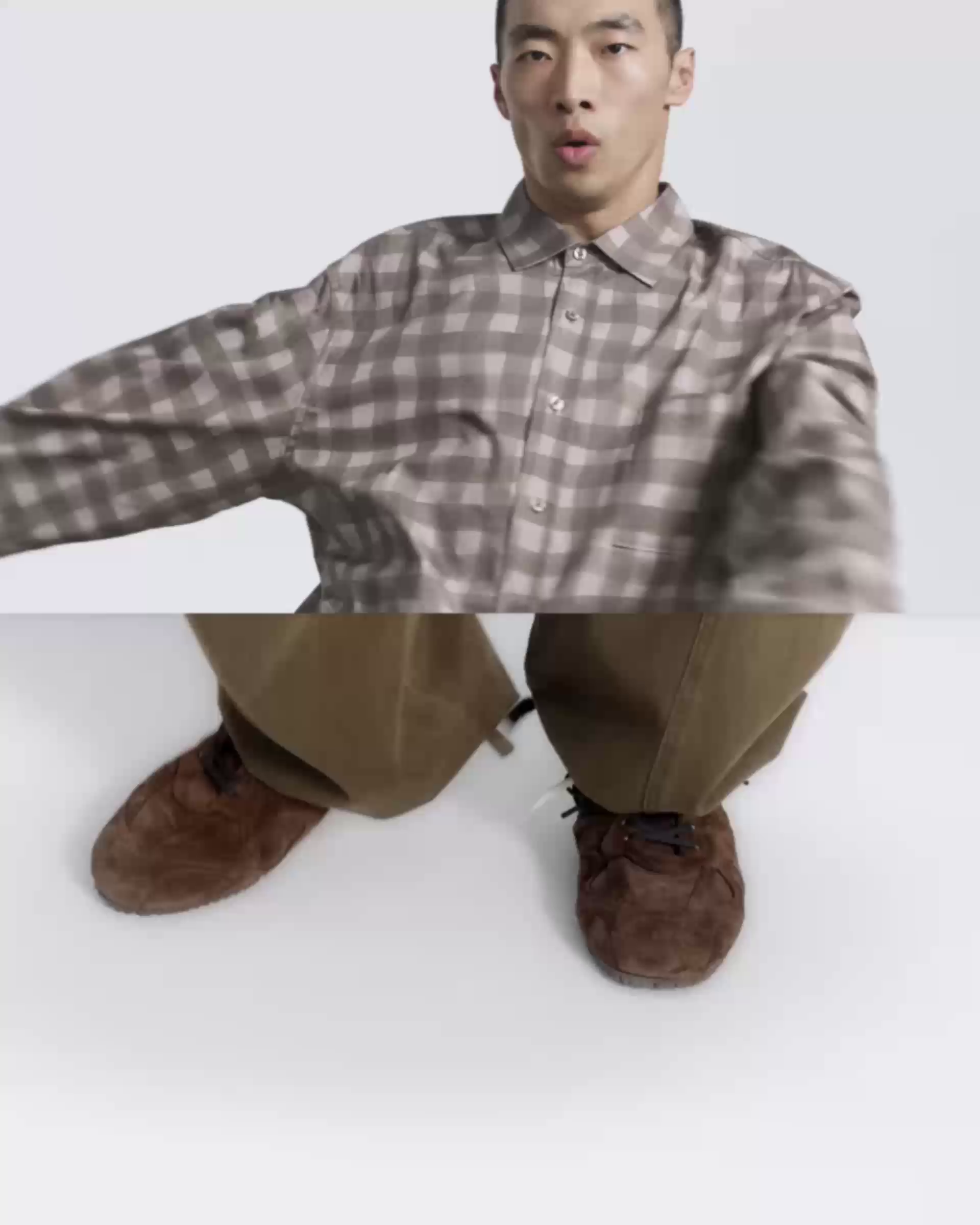 Person wearing a brown plaid shirt and brown pants with brown shoes, standing against a plain background, captured from a low angle.