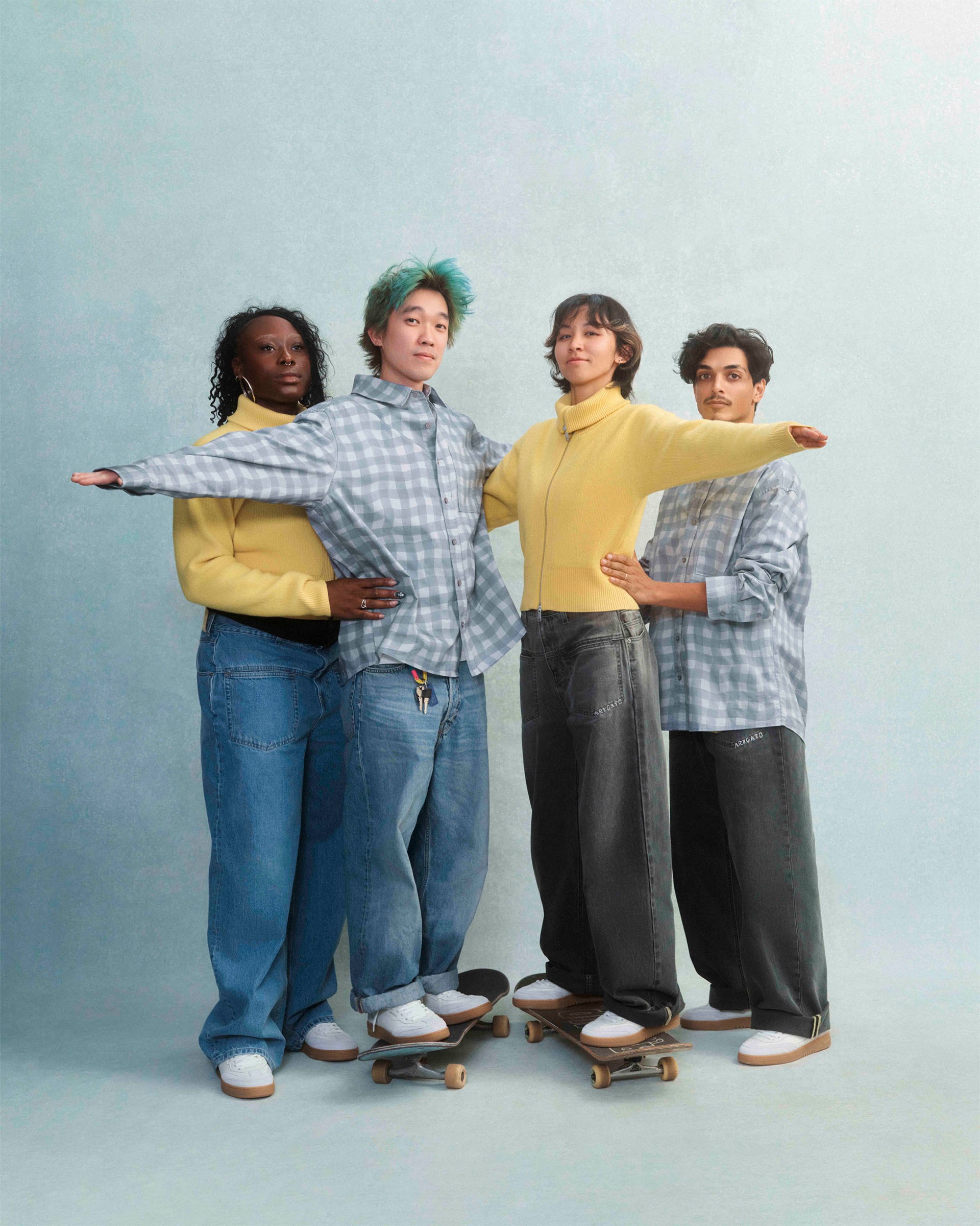 Four people in casual outfits balancing on skateboards, forming a close-knit group against a plain background.