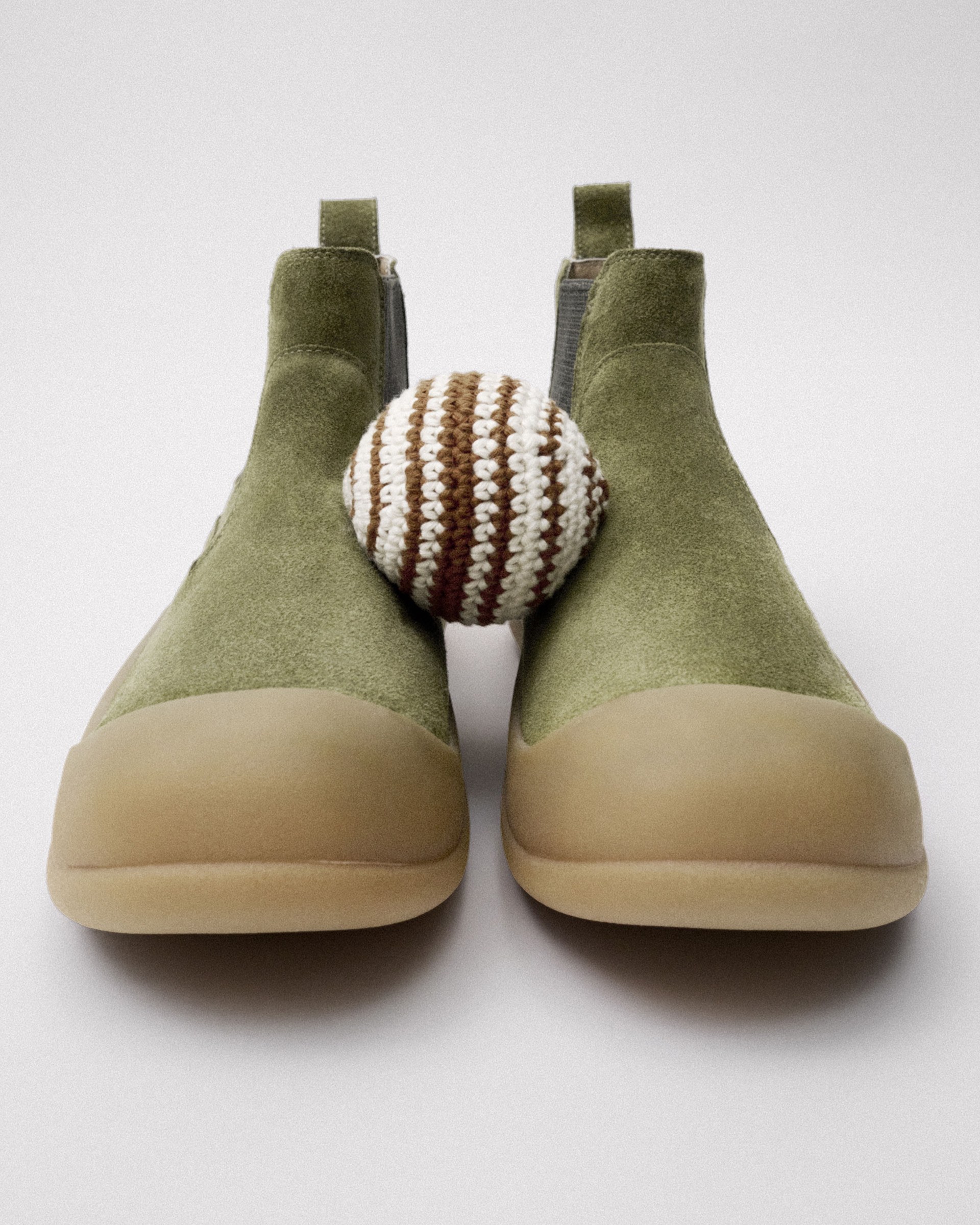 Green suede boots with tan soles, placed side by side, with a small brown and white striped crocheted ball in between them.