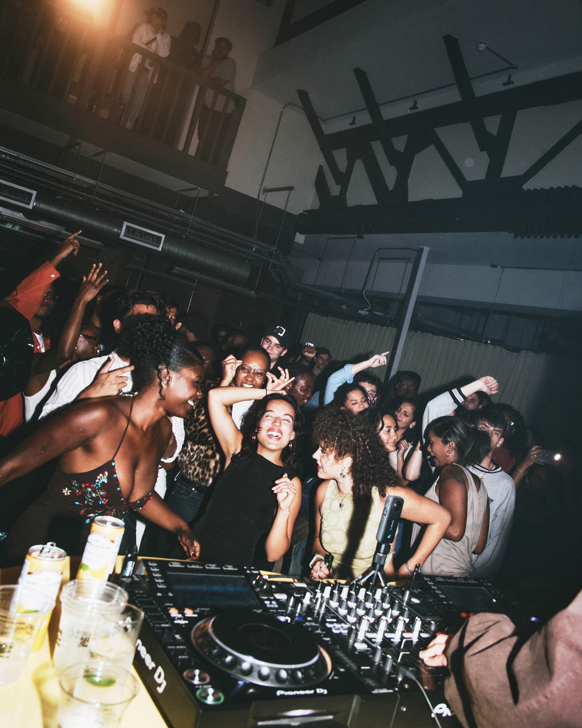 A lively crowd dancing and enjoying music at a club, with DJ equipment visible in the foreground and drinks on a nearby table.