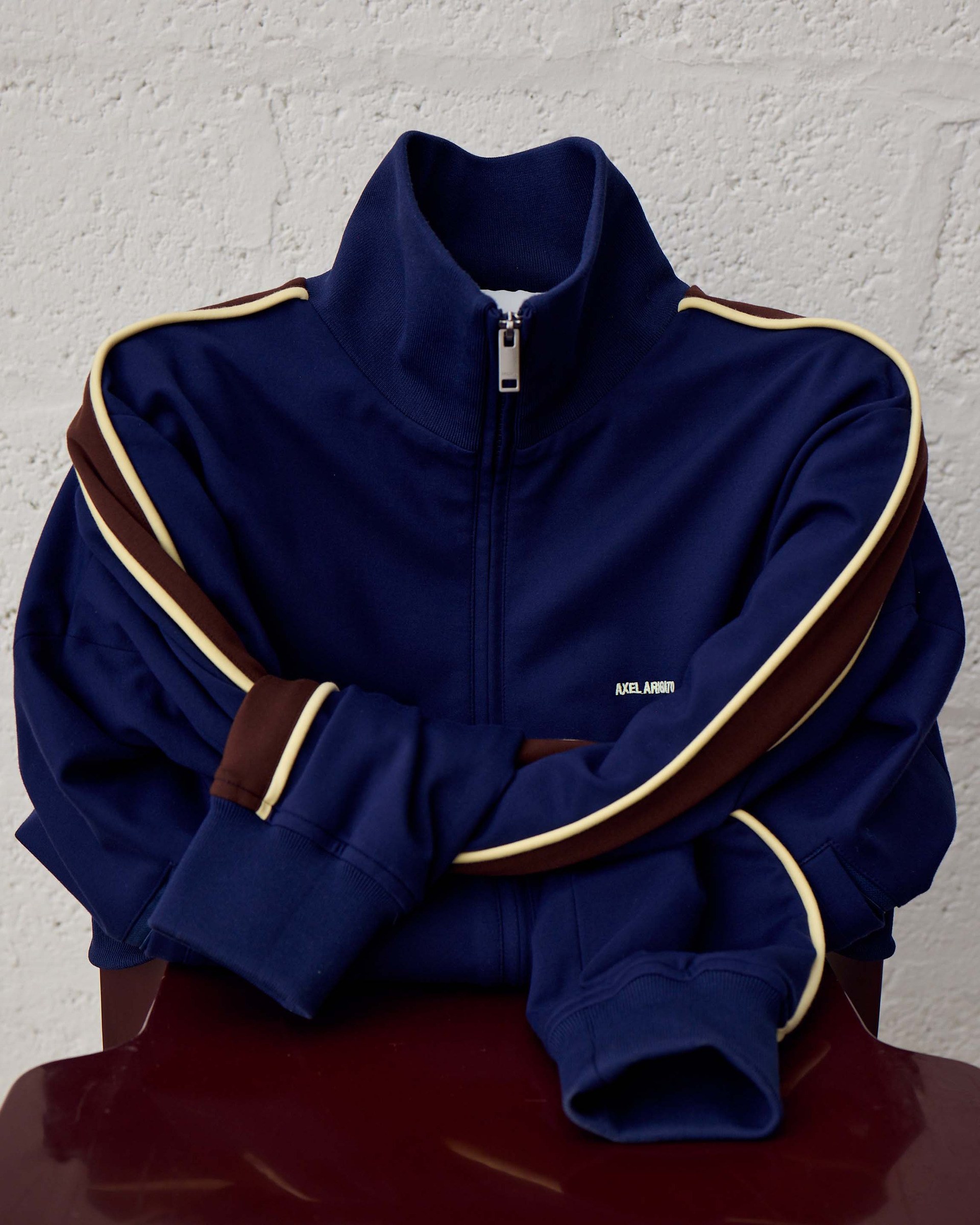 Dark blue zip-up jacket with white and brown stripes on the sleeves, draped over a red chair against a white brick wall.